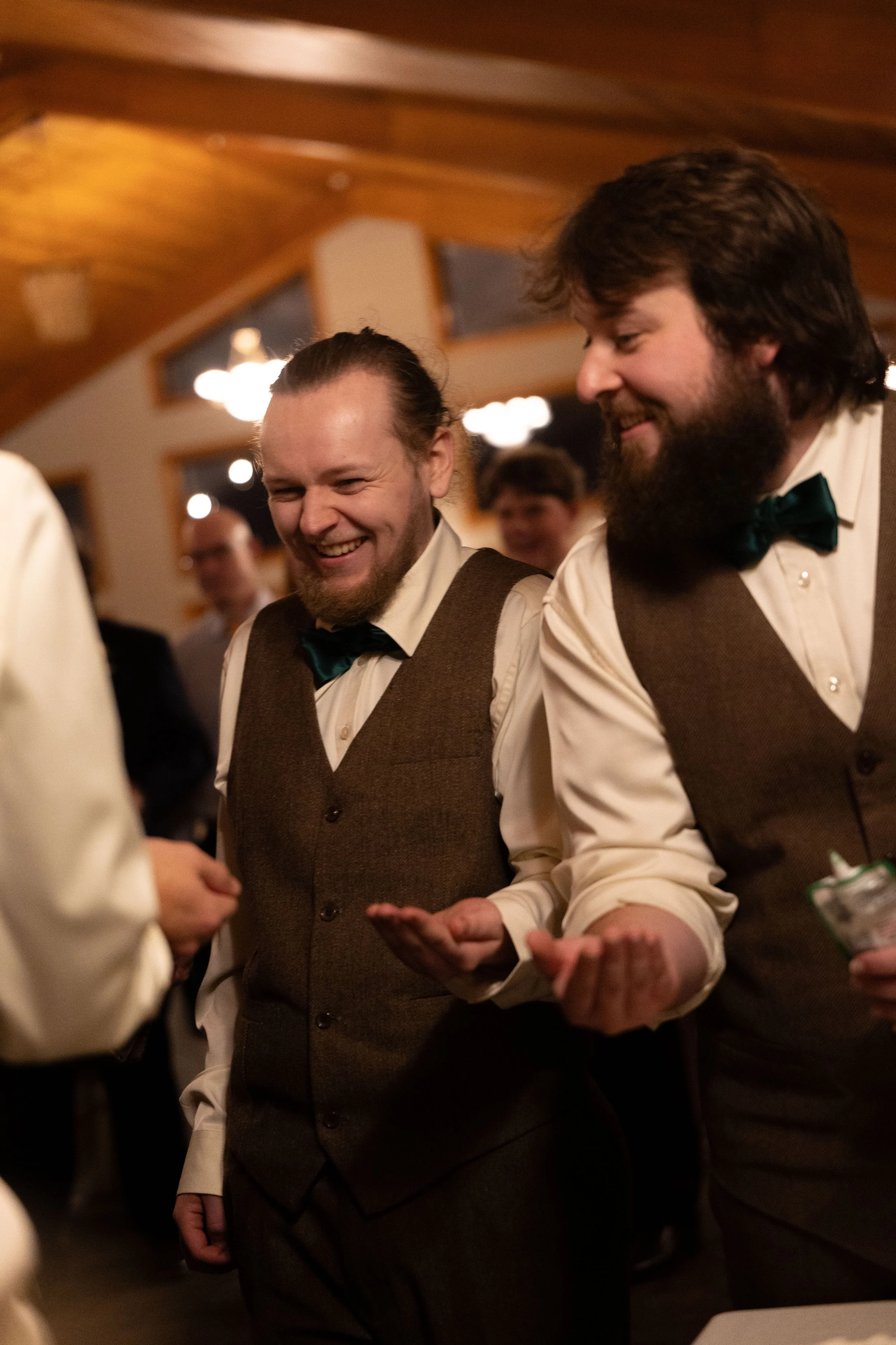 Two men wearing brown vests, white shirts, and black bow ties smile and talk at a social gathering in a warmly lit room.