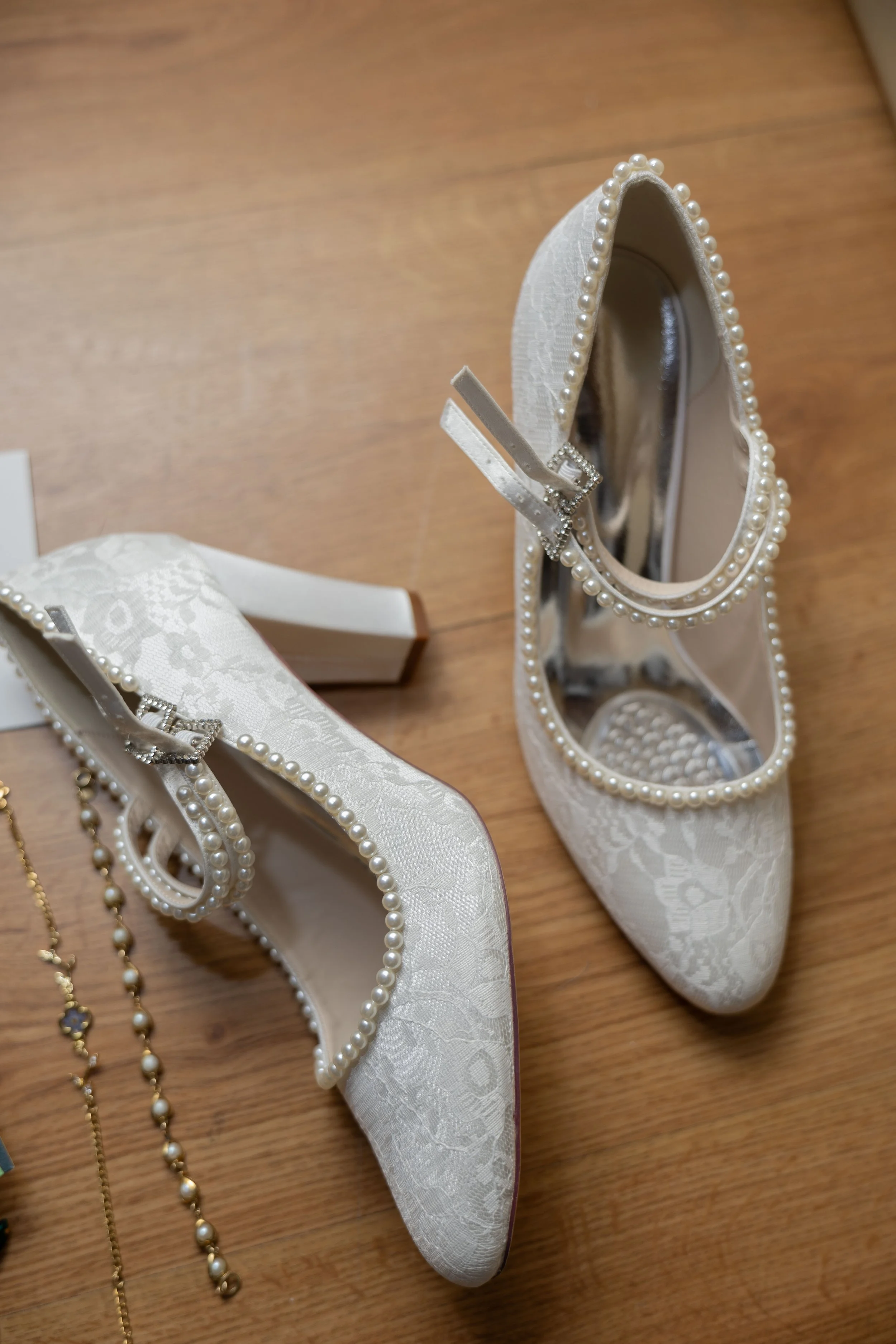 A pair of elegant white lace high-heeled shoes with pearl embellishments and a bow, placed on a wooden surface, with jewelry nearby.