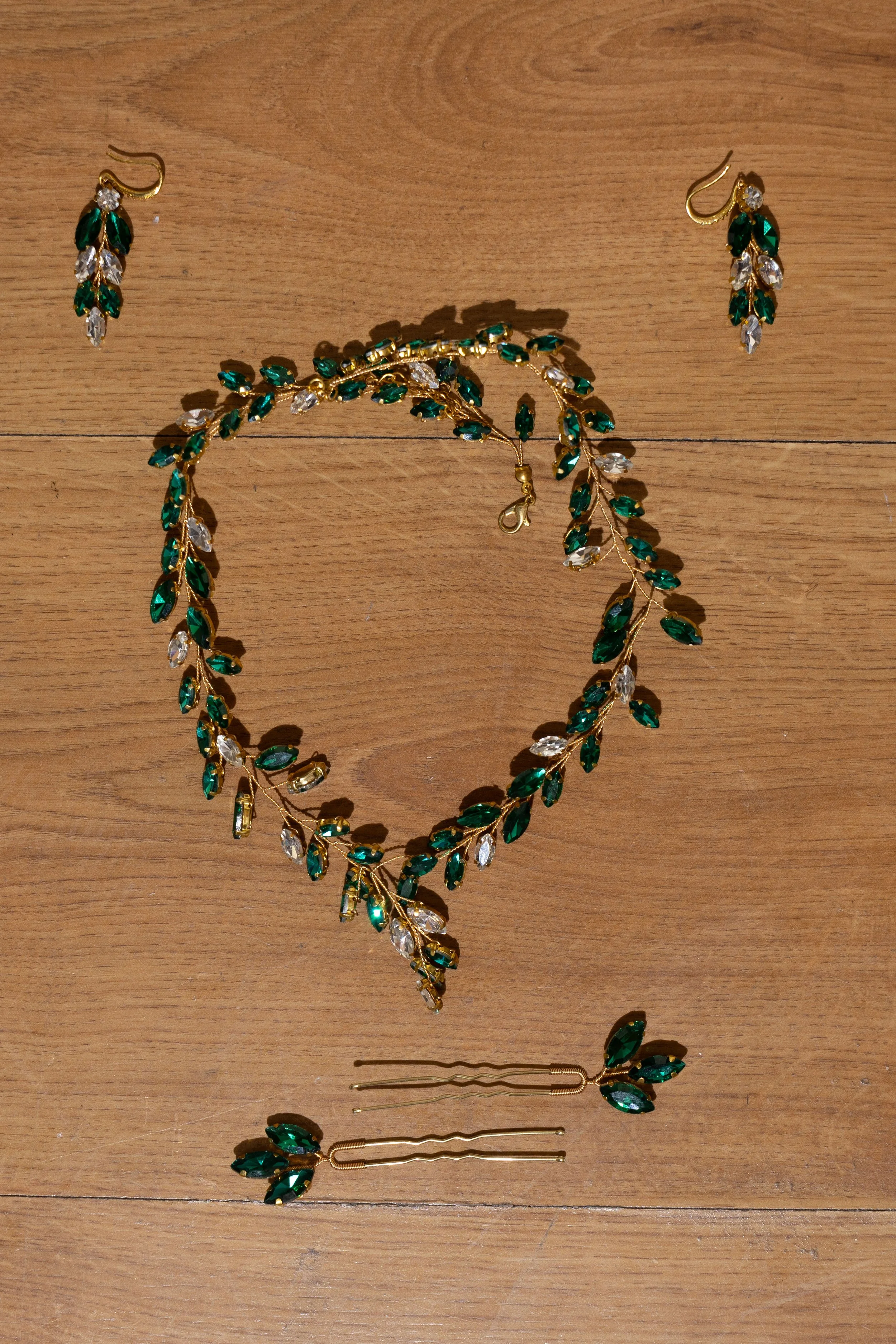 A necklace and matching earrings set with green and clear gemstones arranged in a leaf pattern on a wooden surface.