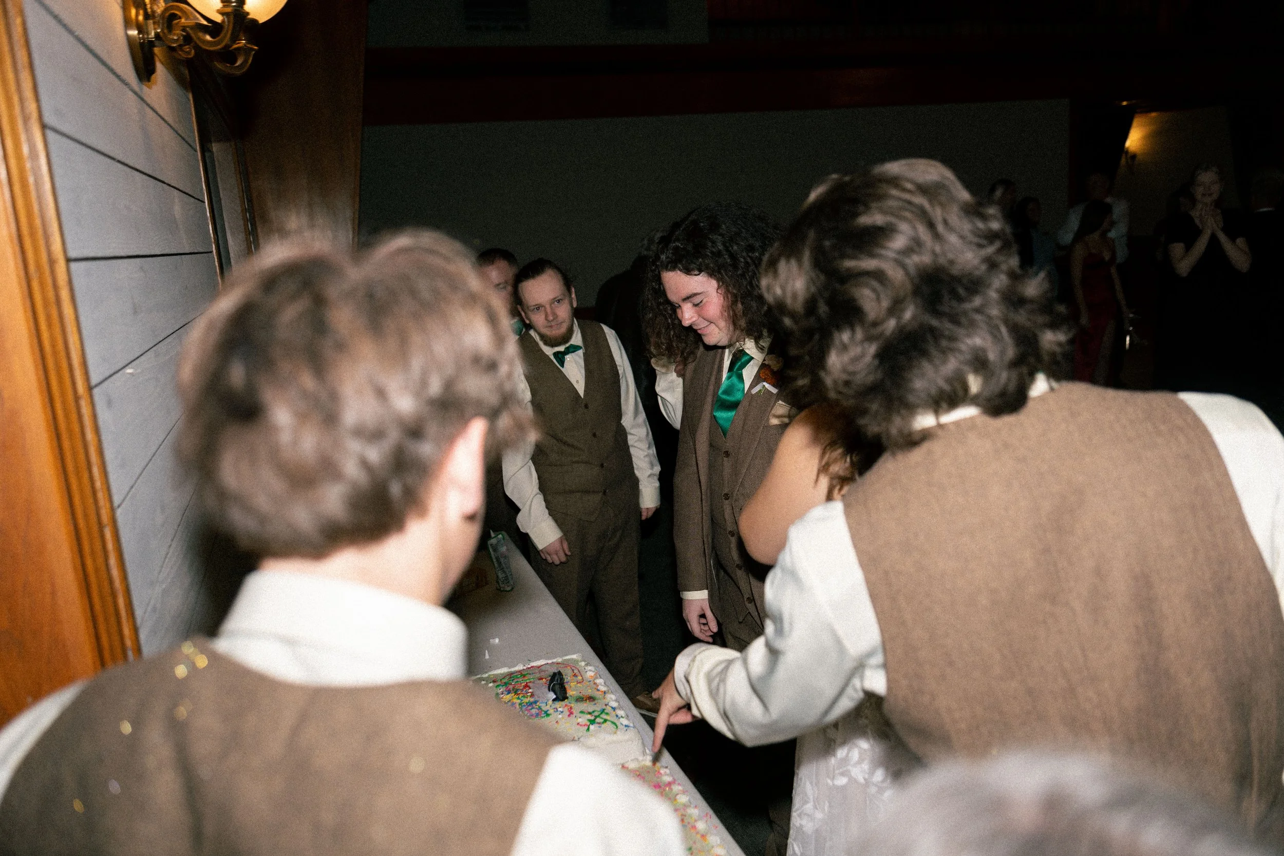 Group of people cutting a decorated birthday cake at a party, with some guests in the background.