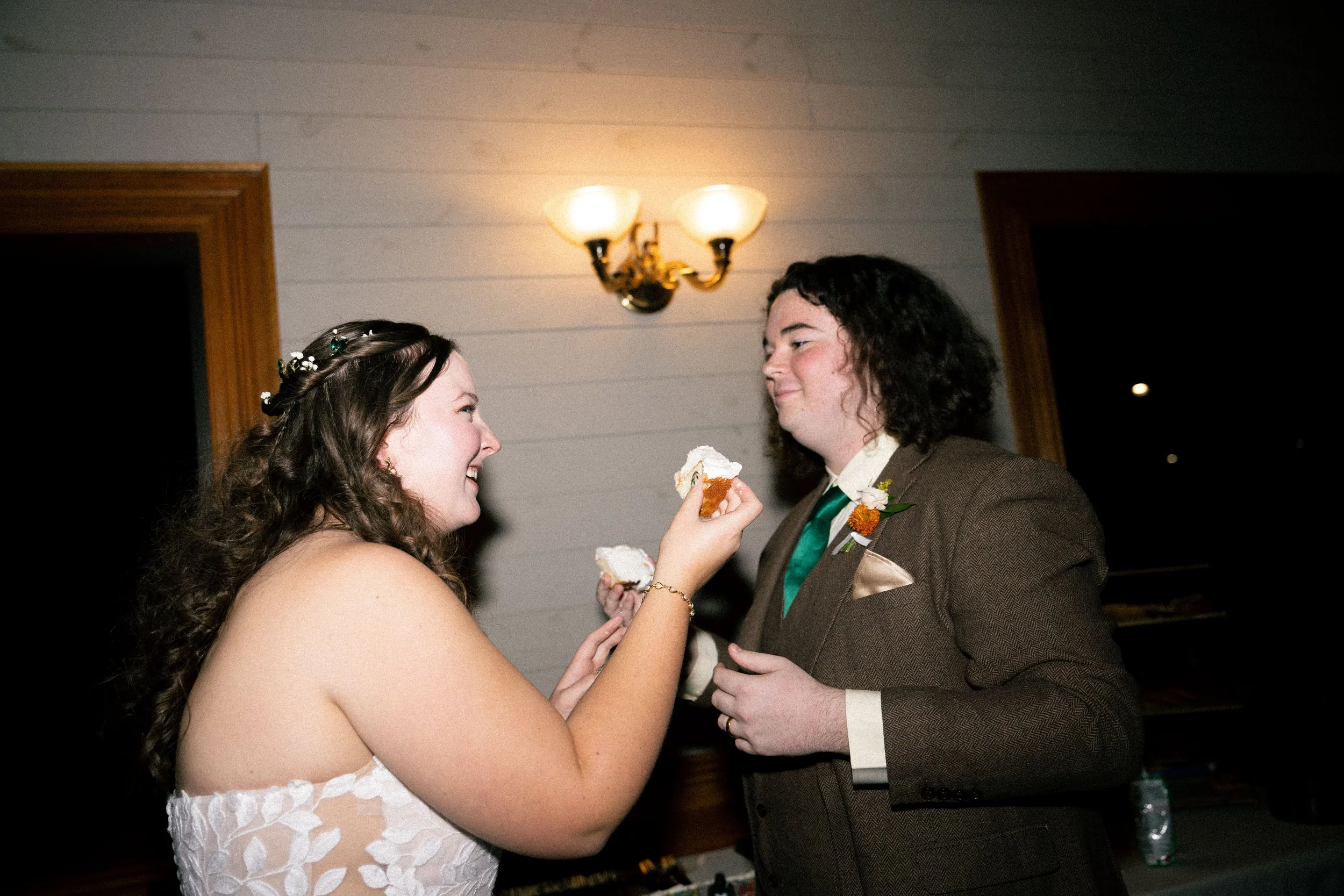 A bride and groom share a moment during their wedding reception. The bride has long curly hair with a floral headpiece and is wearing a white dress, smiling with a piece of cake. The groom has long curly hair, is wearing a brown suit with green tie a