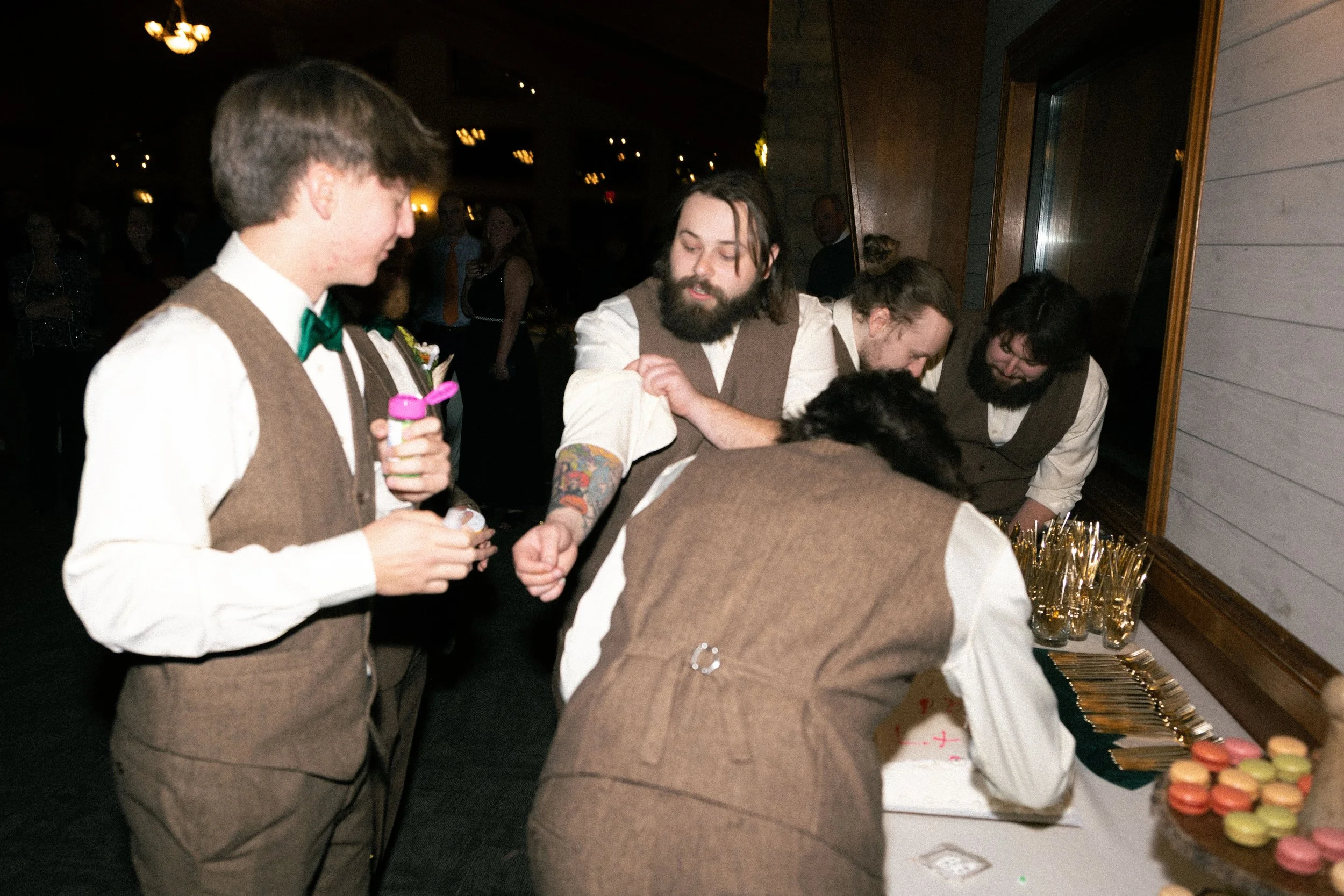 A group of men in vintage-style clothing, gathered around a dessert table, serving macarons at an indoor event.
