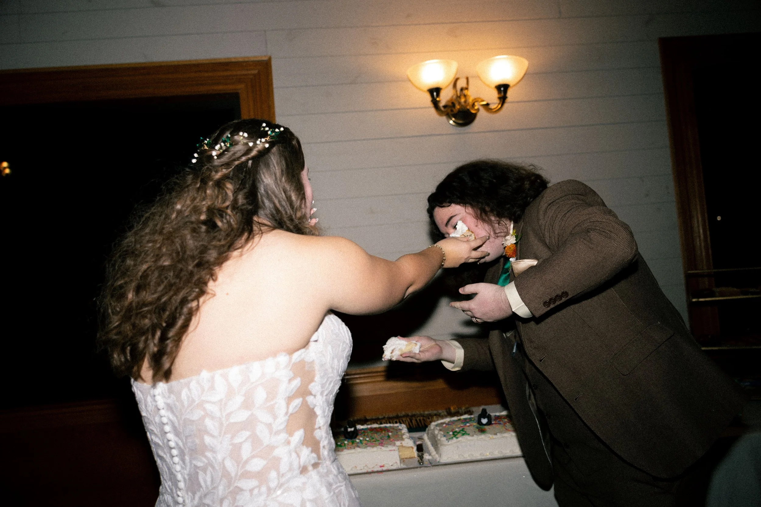 A bride in a lace wedding dress is playfully feeding a piece of cake to a man in a suit during a wedding reception.