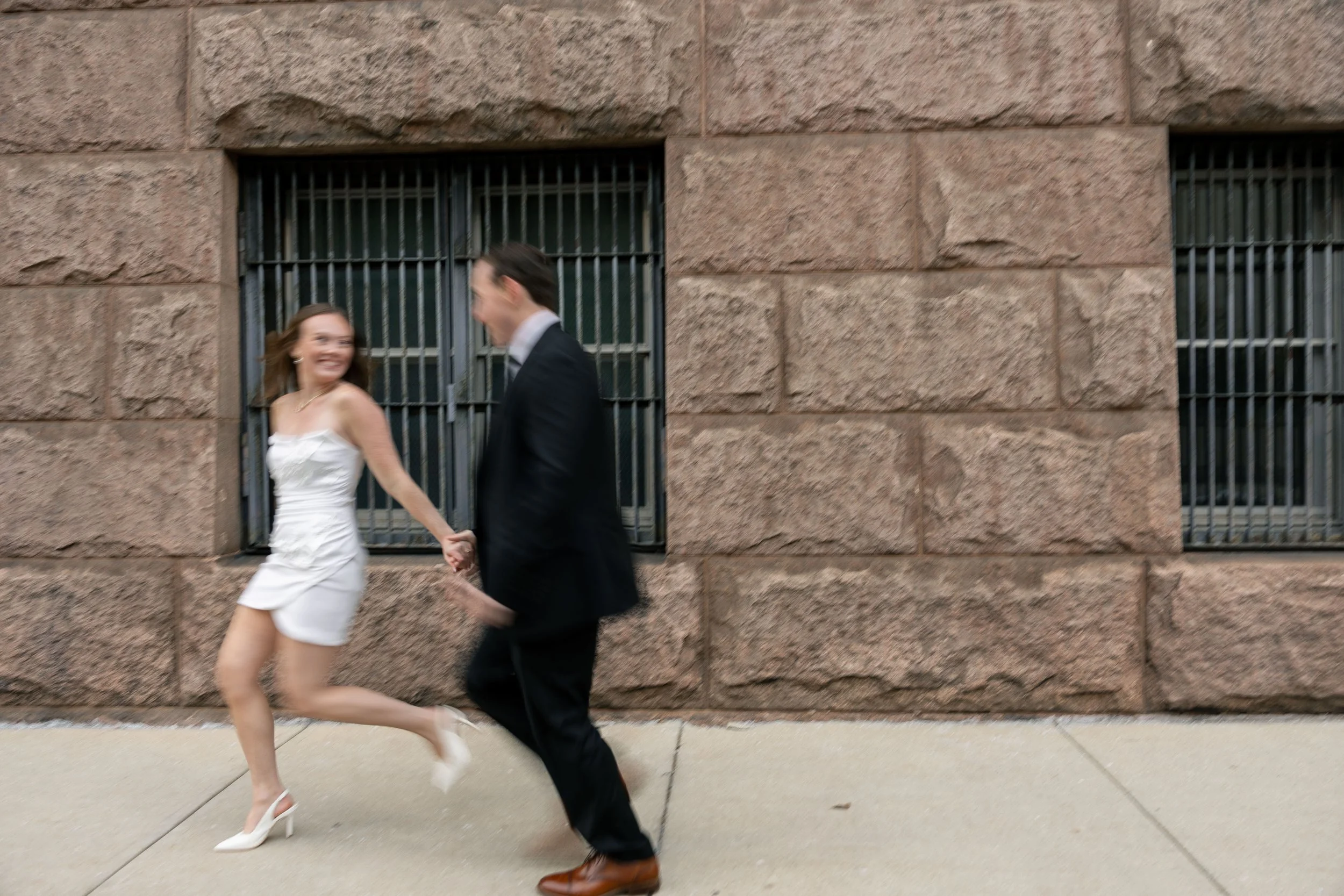 A woman in a white dress and high heels holding hands with a man in a black suit and brown shoes outside a brick building.