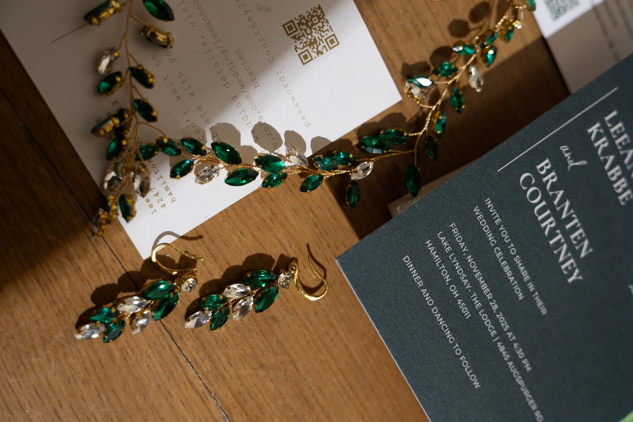 Jewelry set of emerald and clear crystal earrings and a matching necklace alongside a wedding invitation and an RSVP card, arranged on a wooden surface.