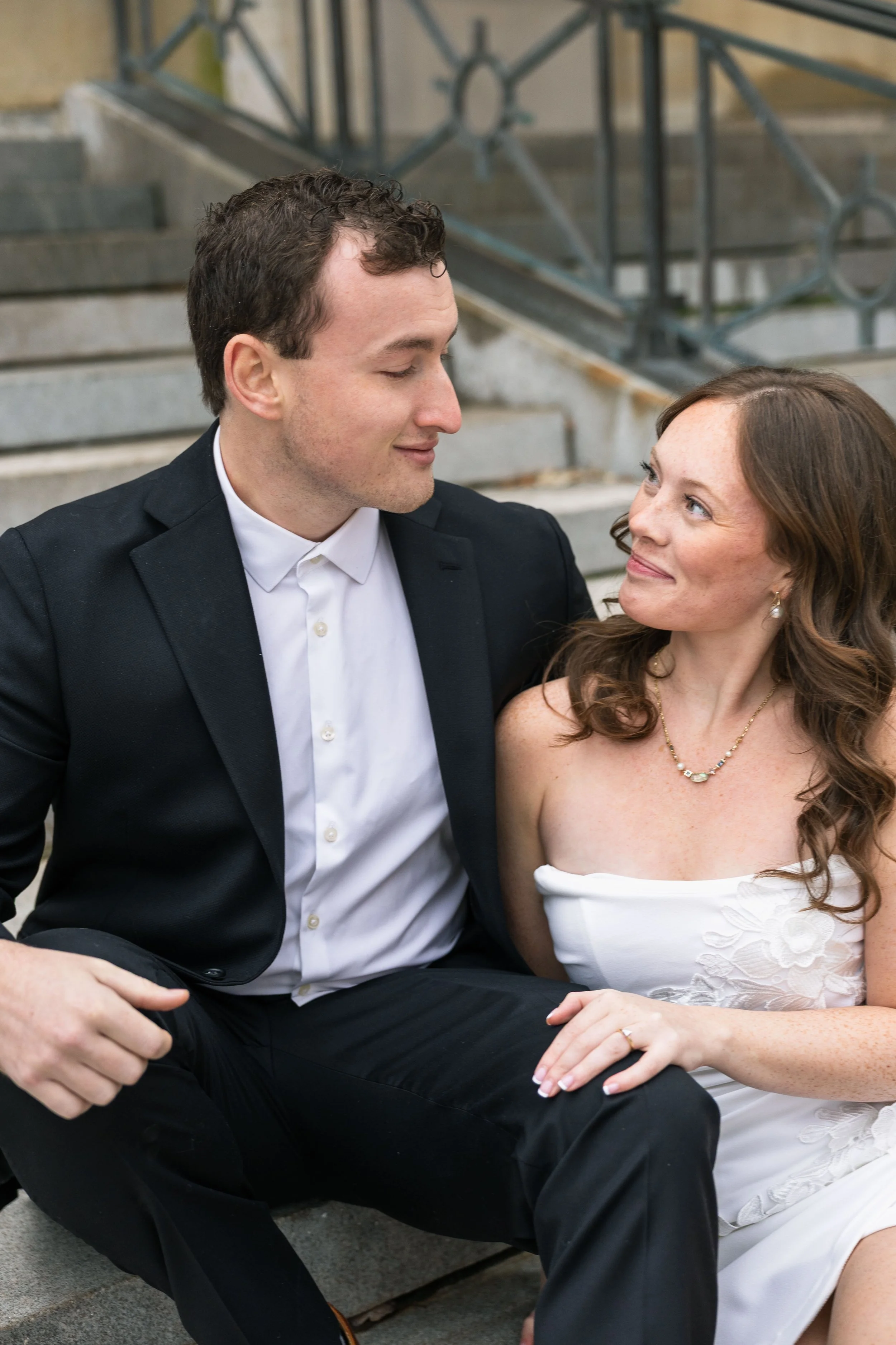 A man and woman sitting on a stairway, looking at each other affectionately. The man is in a black suit with a white shirt, and the woman is wearing a white strapless dress with floral details and jewelry.