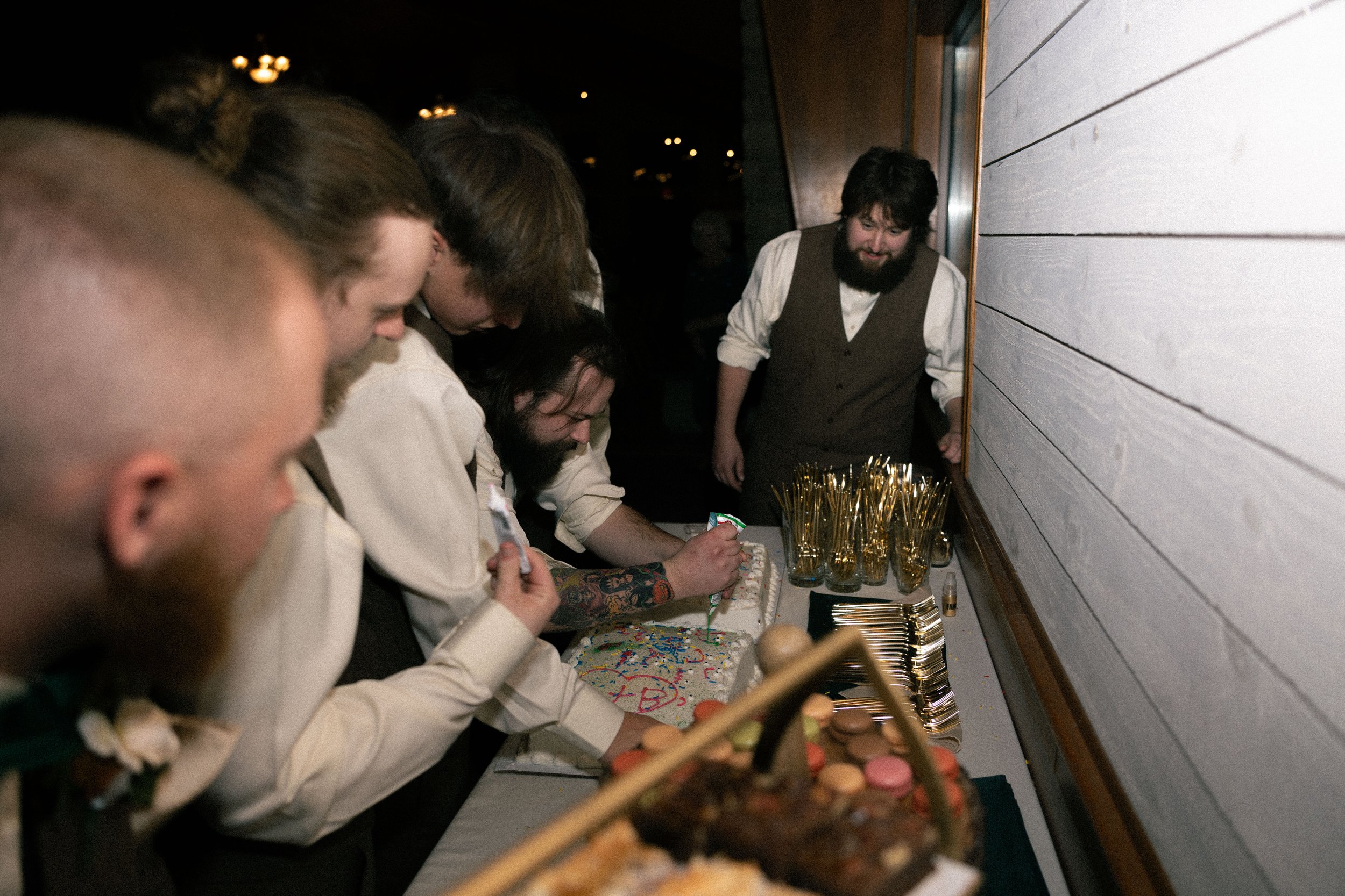 A group of men in vintage clothing gathered around a table with cakes and desserts, with one man writing on a cake.