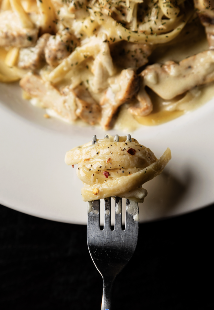 Close-up of a fork holding a piece of cheesy chicken alfredo pasta with red pepper flakes and seasoning, with a plate of pasta in the background.