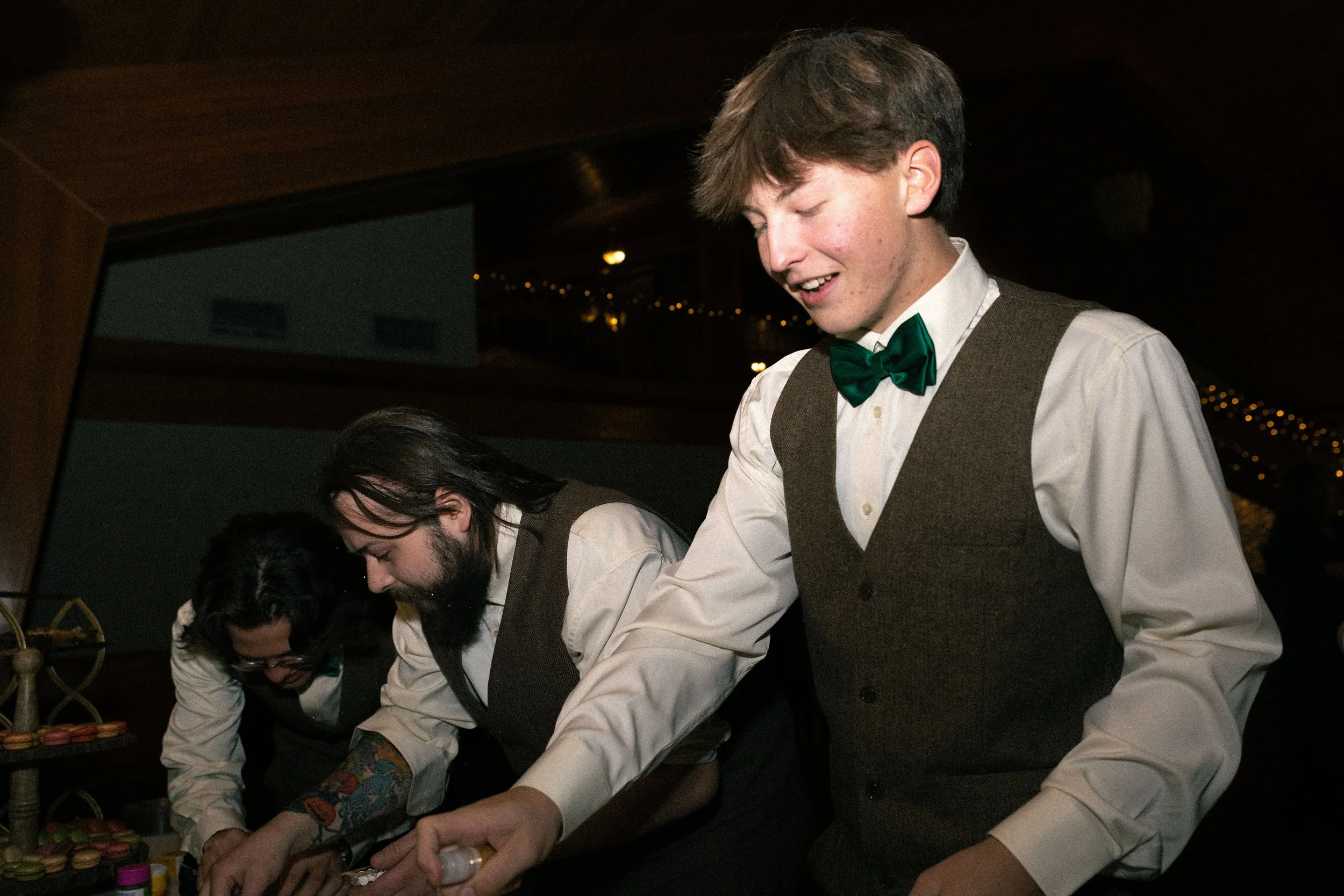 Three people dressed in formal attire, wearing vests and bow ties, appear to be preparing for a food or drink event, with one person smiling and two others focused on their tasks.