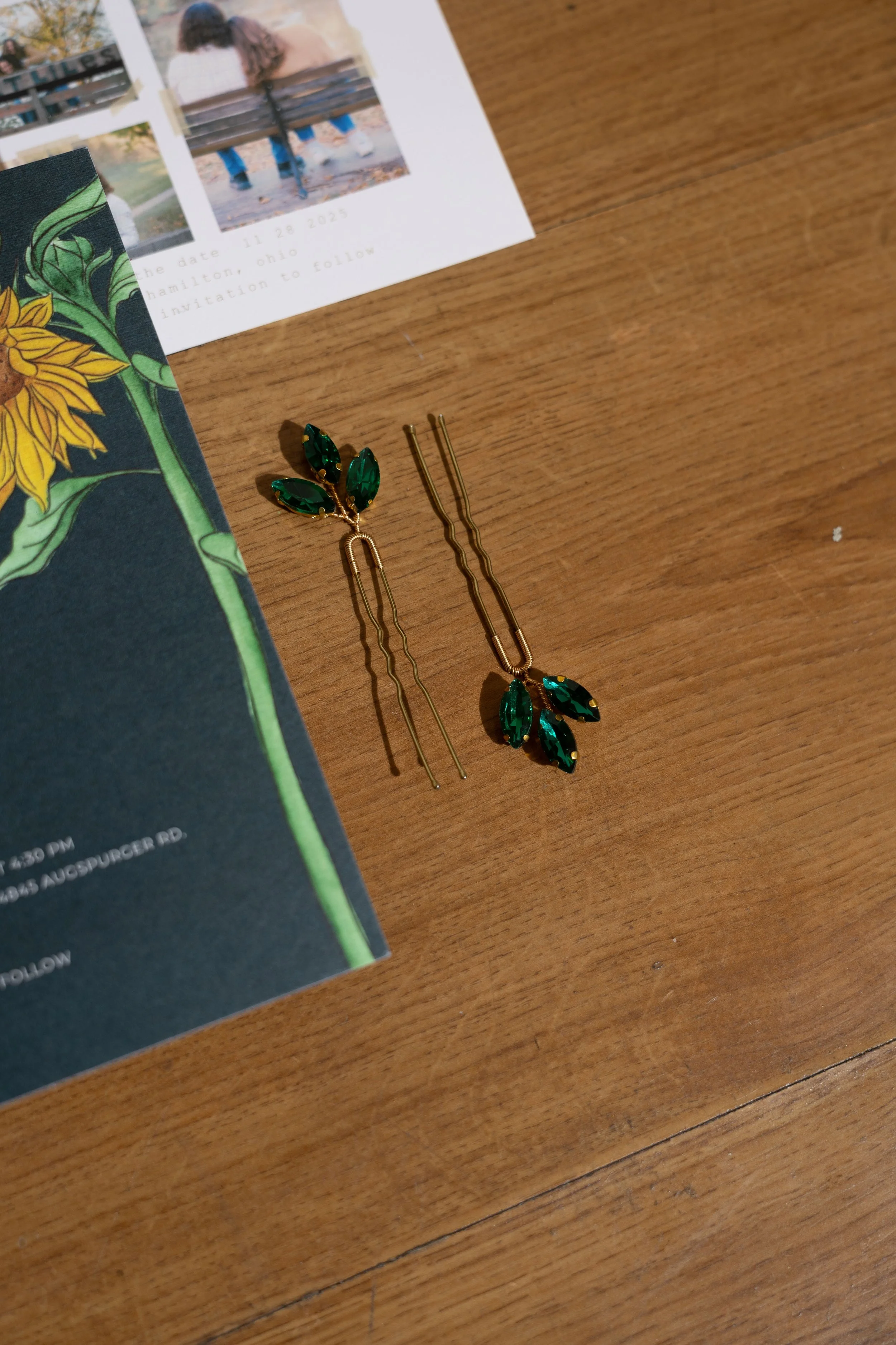Pair of gold-tone earrings with green leaf-shaped stones on a wooden surface, next to a black and green floral-patterned card and a partially visible white paper with a photo of a couple sitting on a bench.