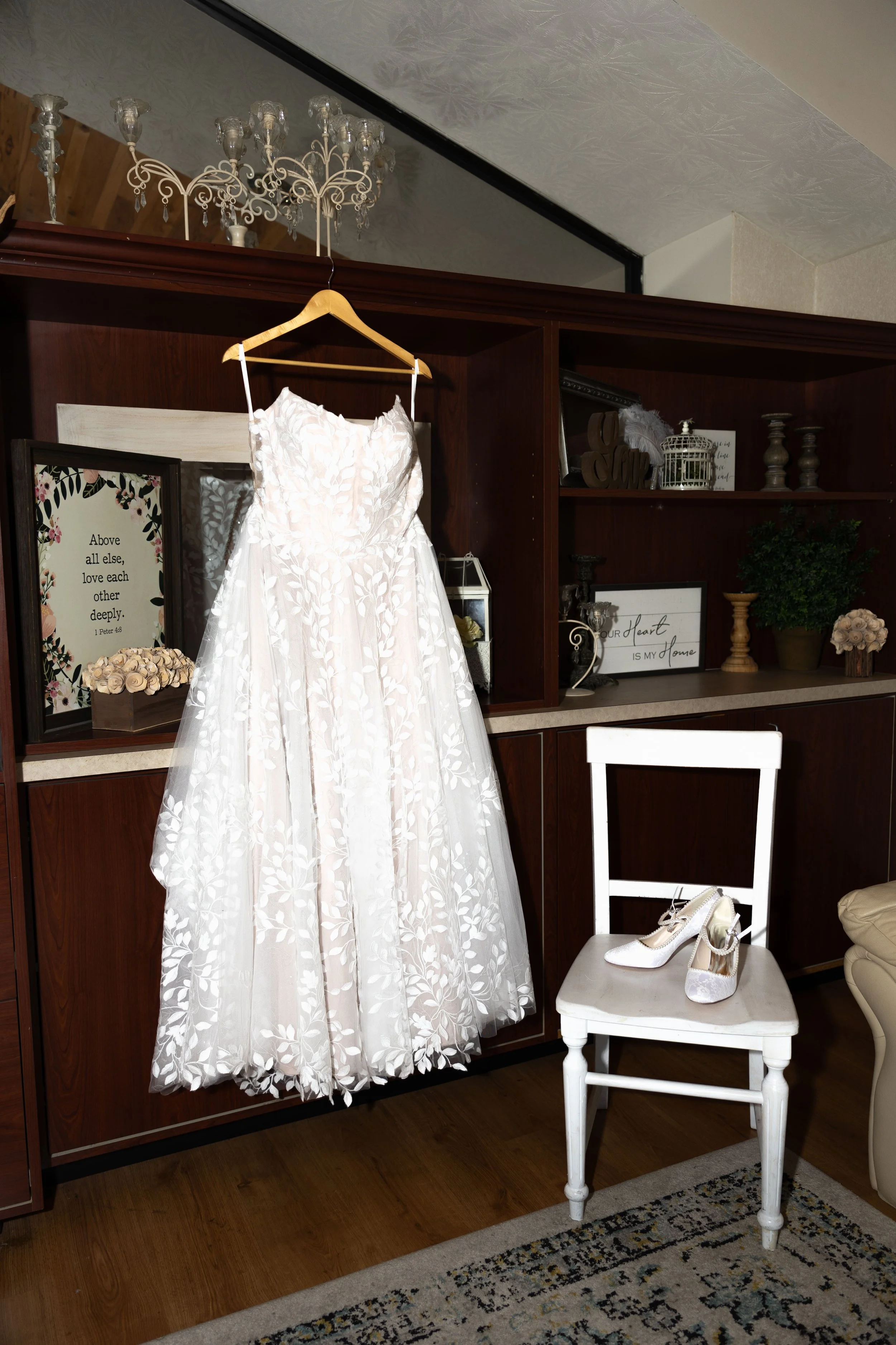 A white wedding dress hanging on a wooden hanger on a dark wood shelf, with white high-heeled shoes on a white chair below it. Decorative items and framed quotes are on the shelf behind, including a sign that reads, ‘Above all else, love each other d