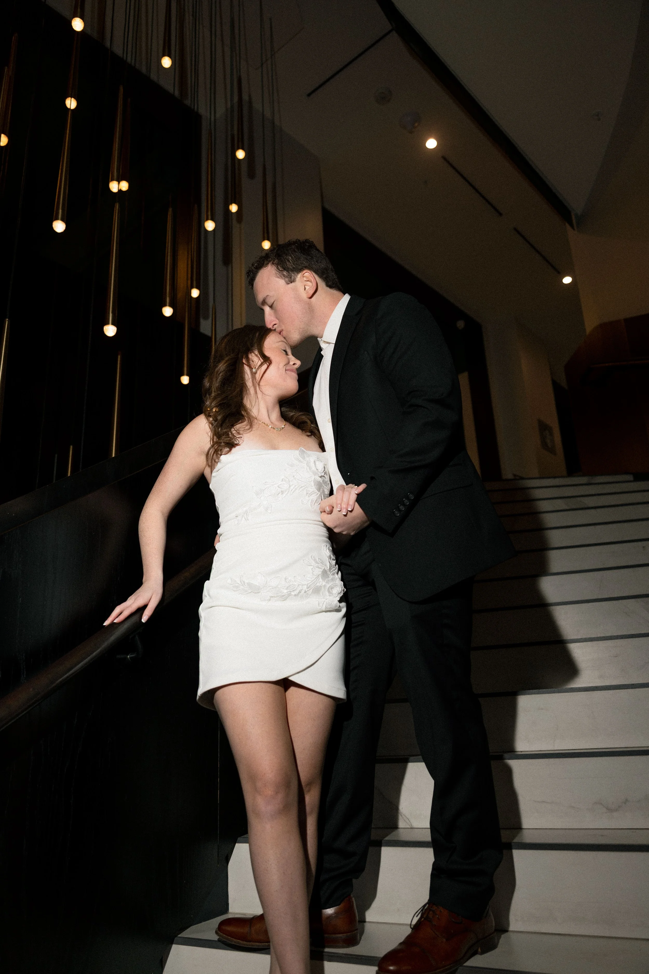 A young couple sharing a romantic moment on a staircase, with the man about to kiss the woman's forehead, in an upscale indoor setting with modern lighting.