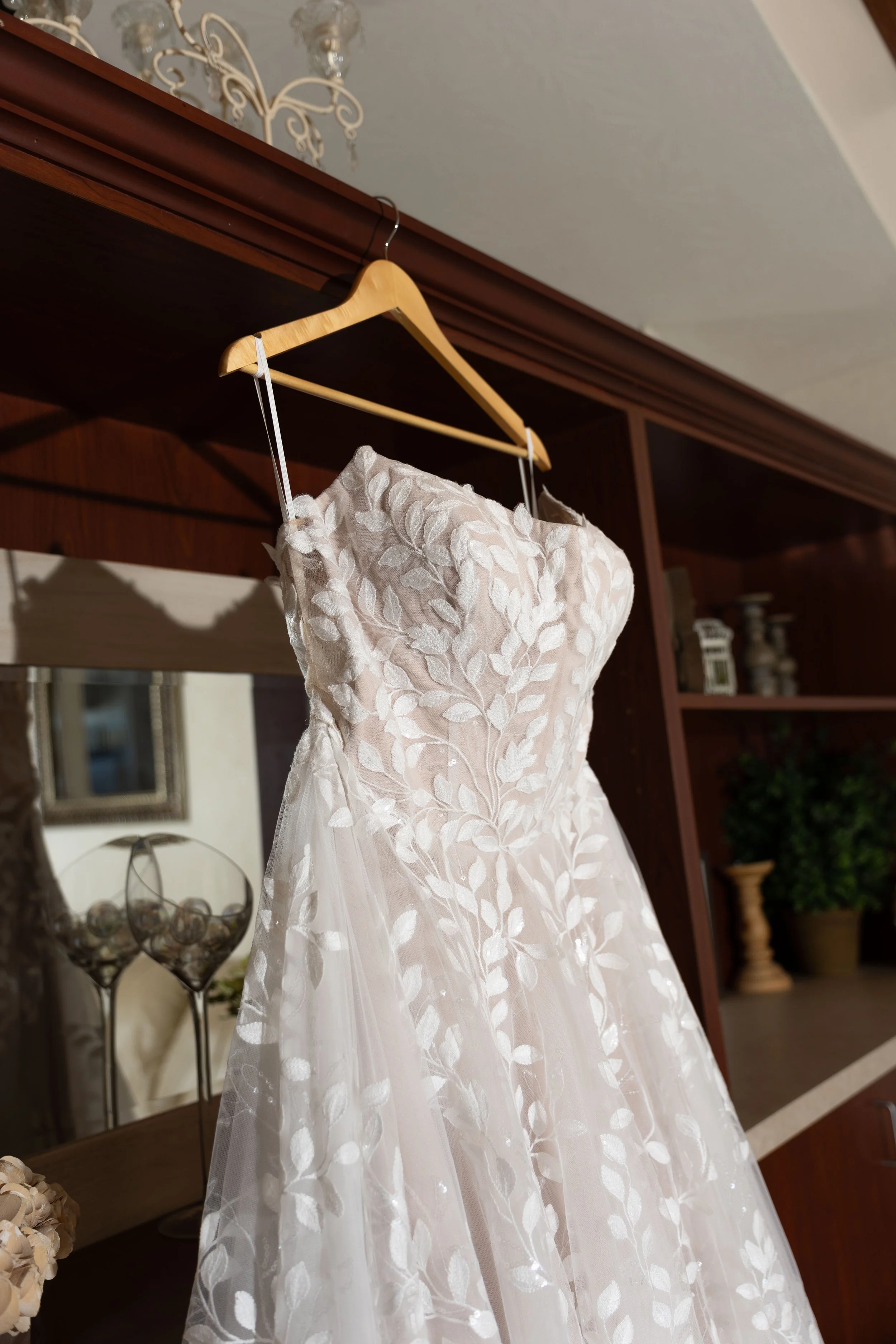 A white wedding dress with lace floral embroidery hanging on a wooden hanger in a room with wooden furniture and decorative items.
