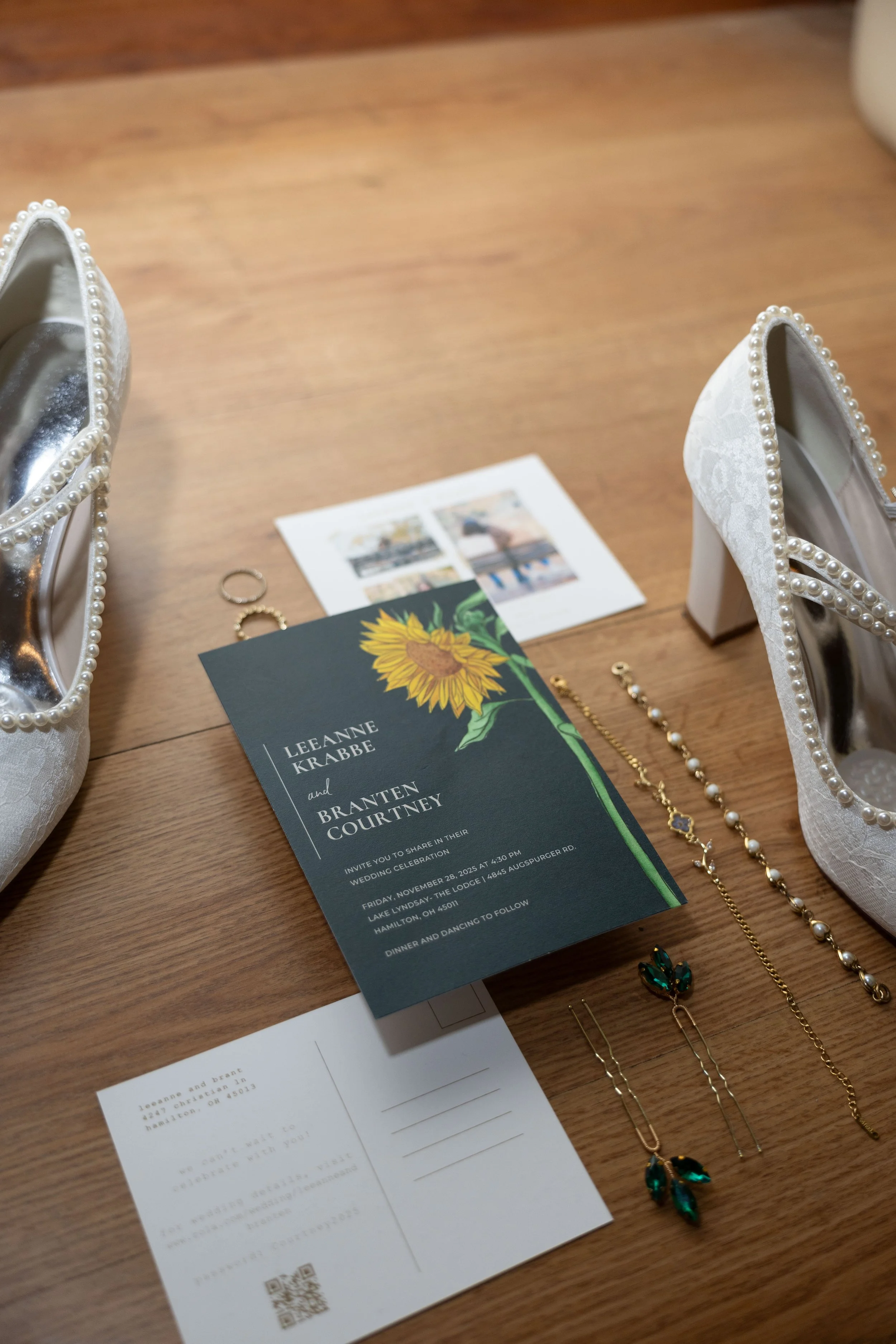 Wedding invitation, pearl decorated white shoes, jewelry, and a thank you card on a wooden table.