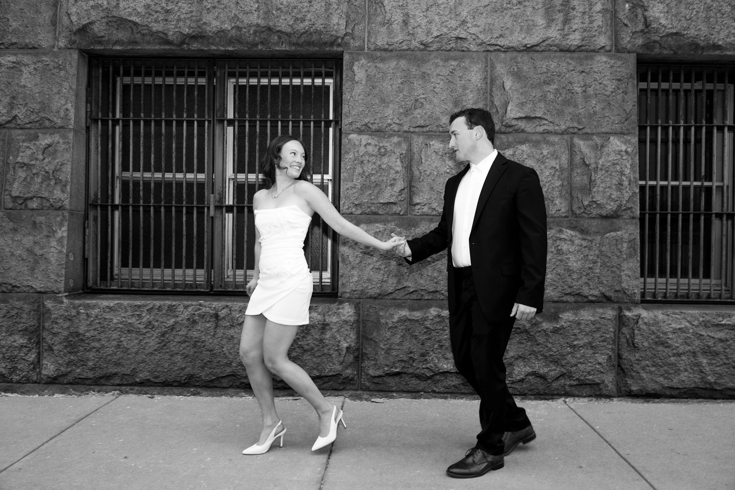 A black-and-white photo of a woman in a white dress and high heels, and a man in a suit, holding hands and smiling at each other on a city sidewalk in front of a stone building with barred windows.