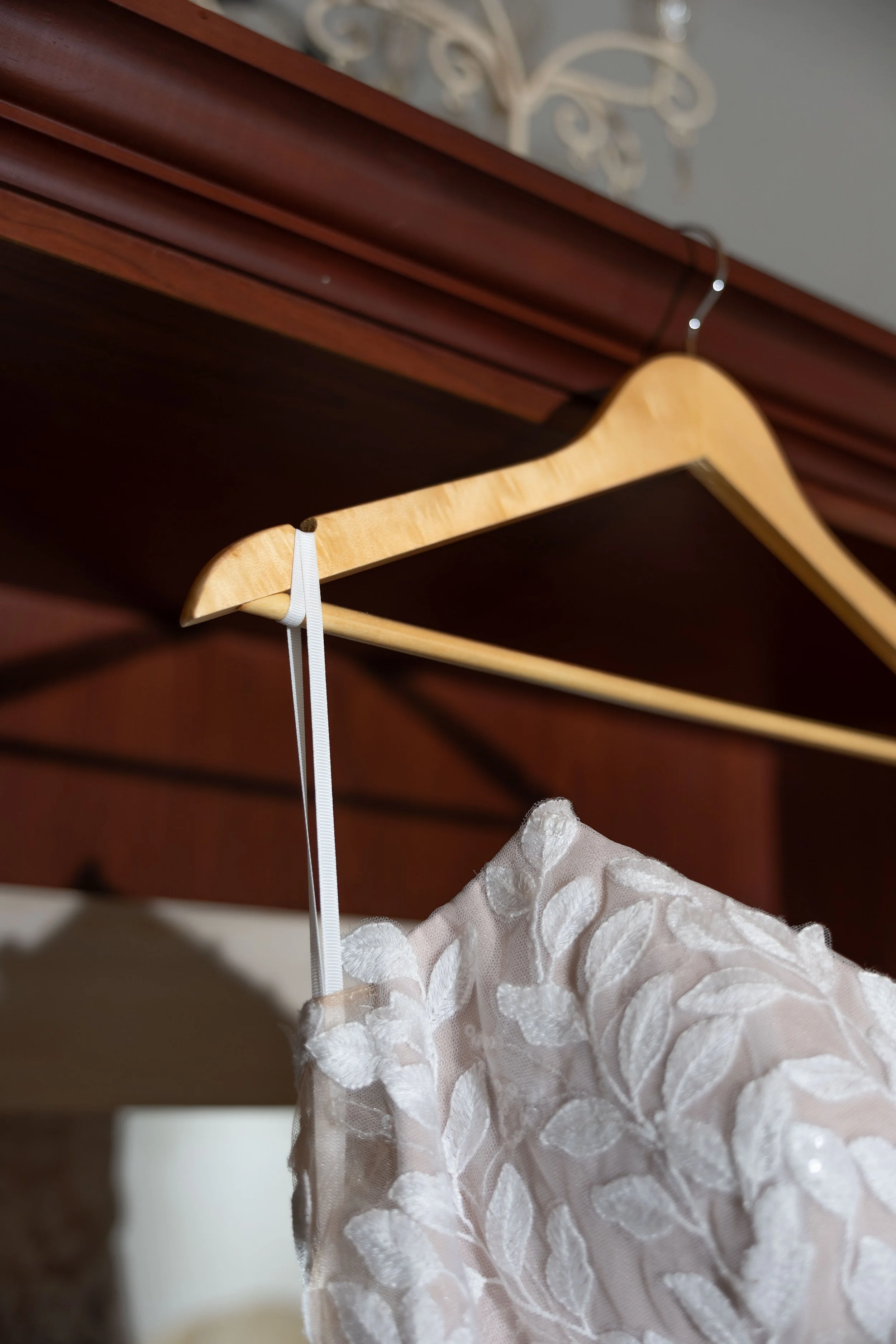 Close-up of a white wedding dress with floral embroidery hanging on a wooden hanger in a closet.