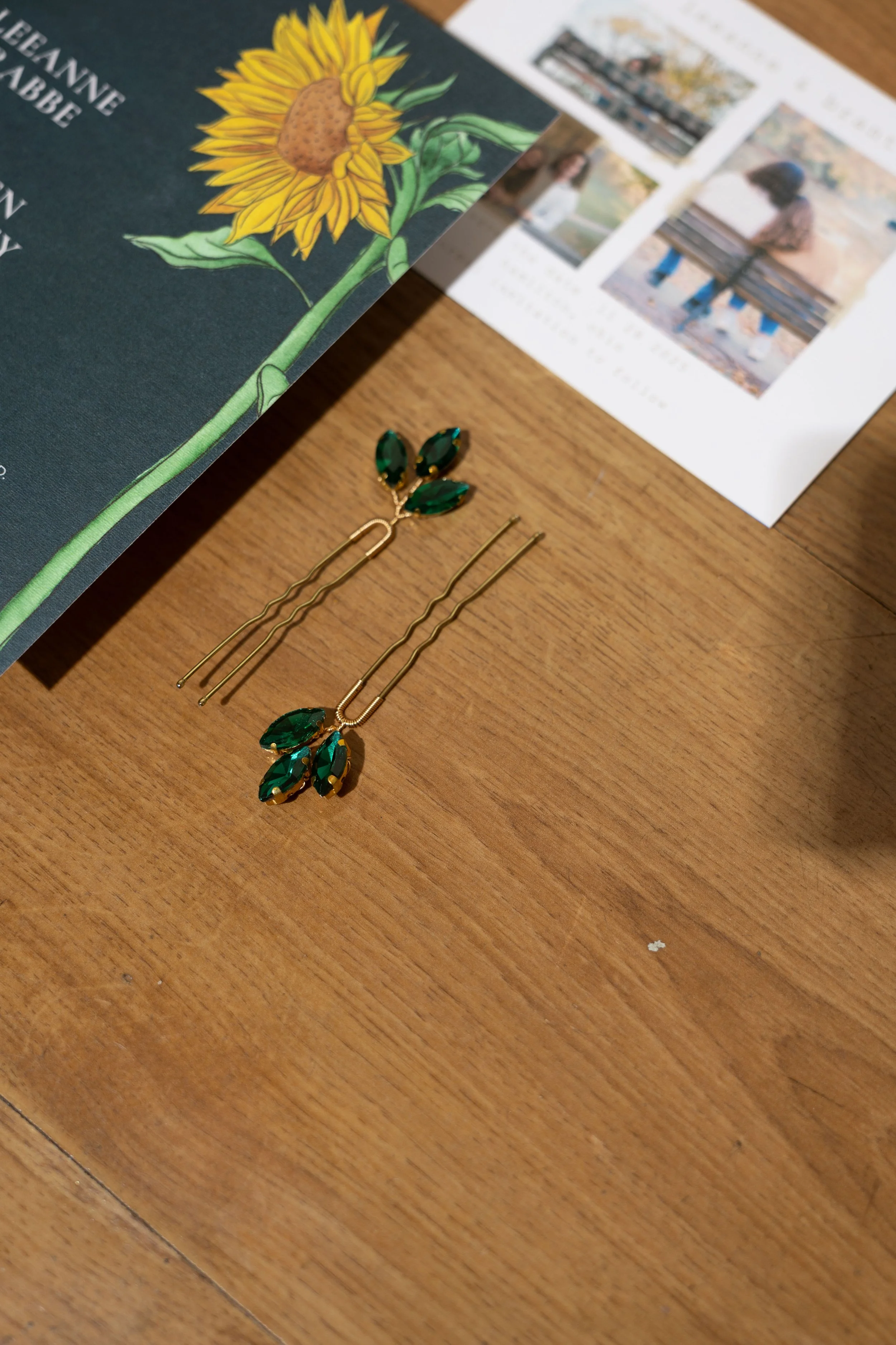Green and gold decorative hairpins with leaf design on a wooden table near a magazine with a sunflower illustration and some photos.