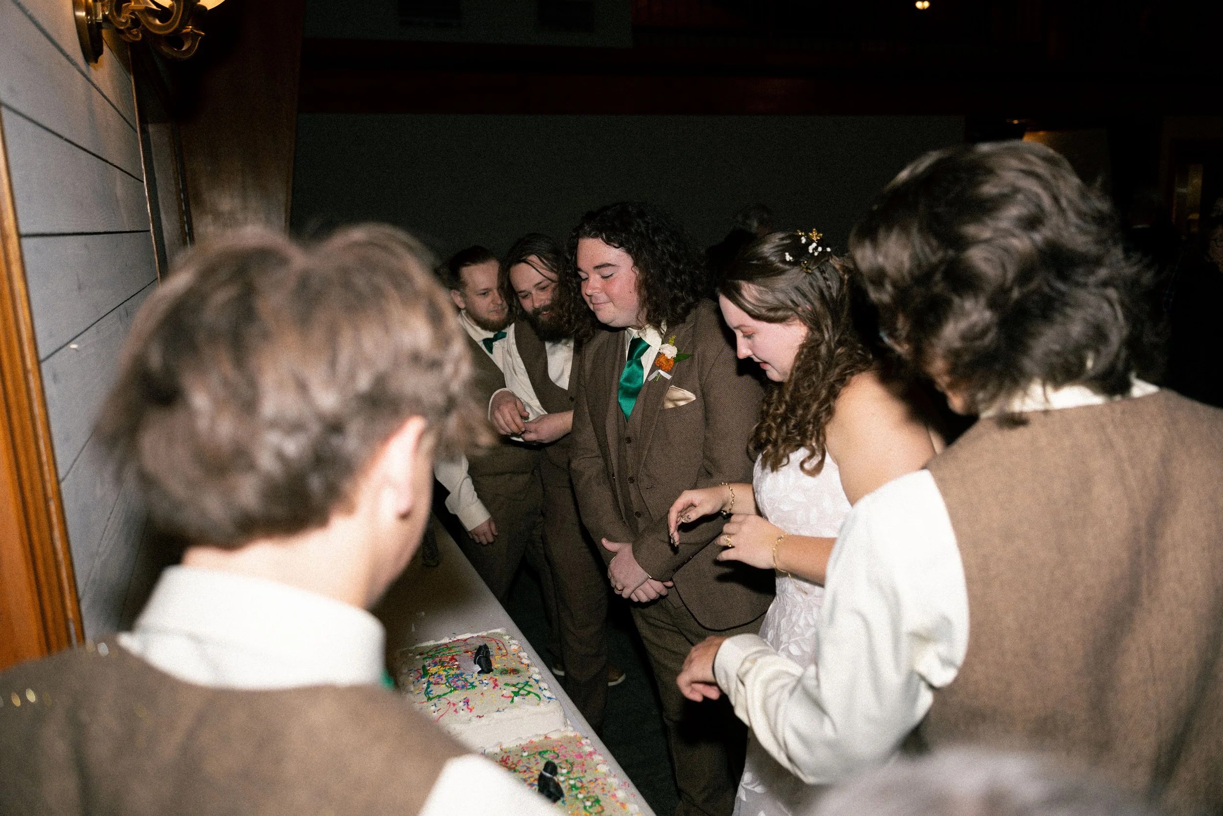 People gathered around a table with a decorated cake, celebrating a wedding or special occasion, with a woman in a white dress and others in formal attire.