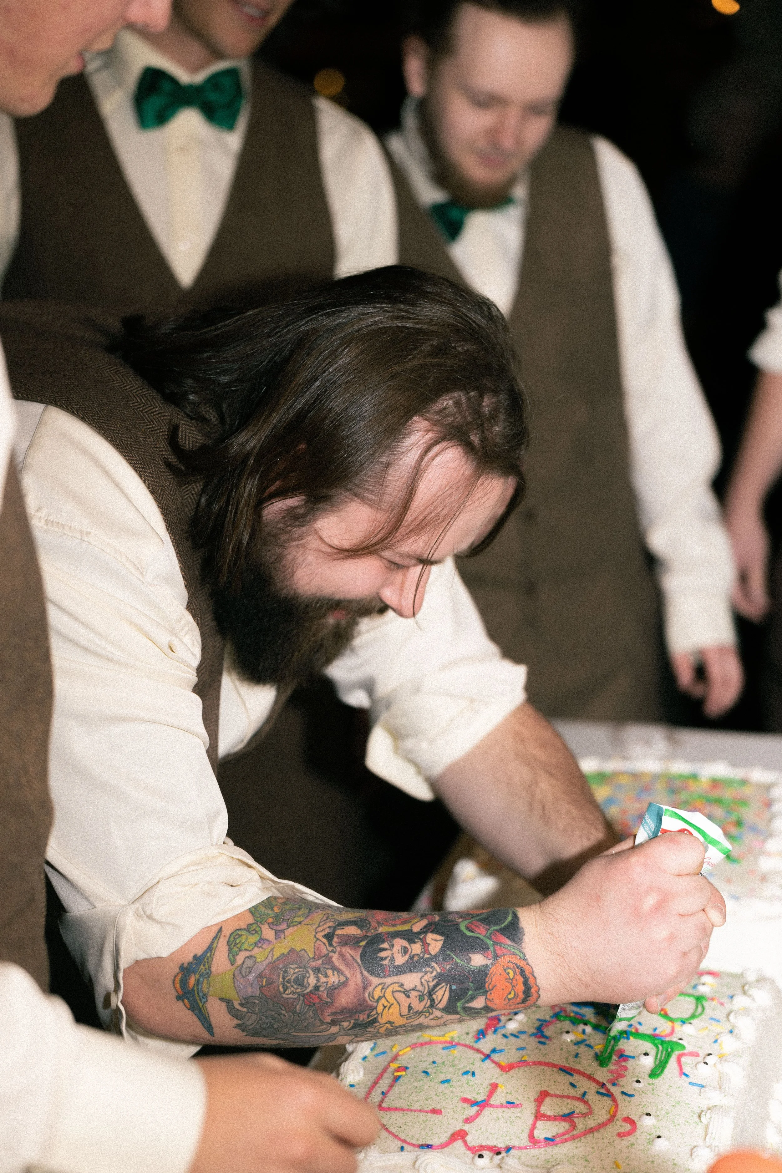 A man with long hair and tattoos writing on a decorated birthday cake with colorful icing and sprinkles, while others look on. The setting appears to be a celebration or birthday party.