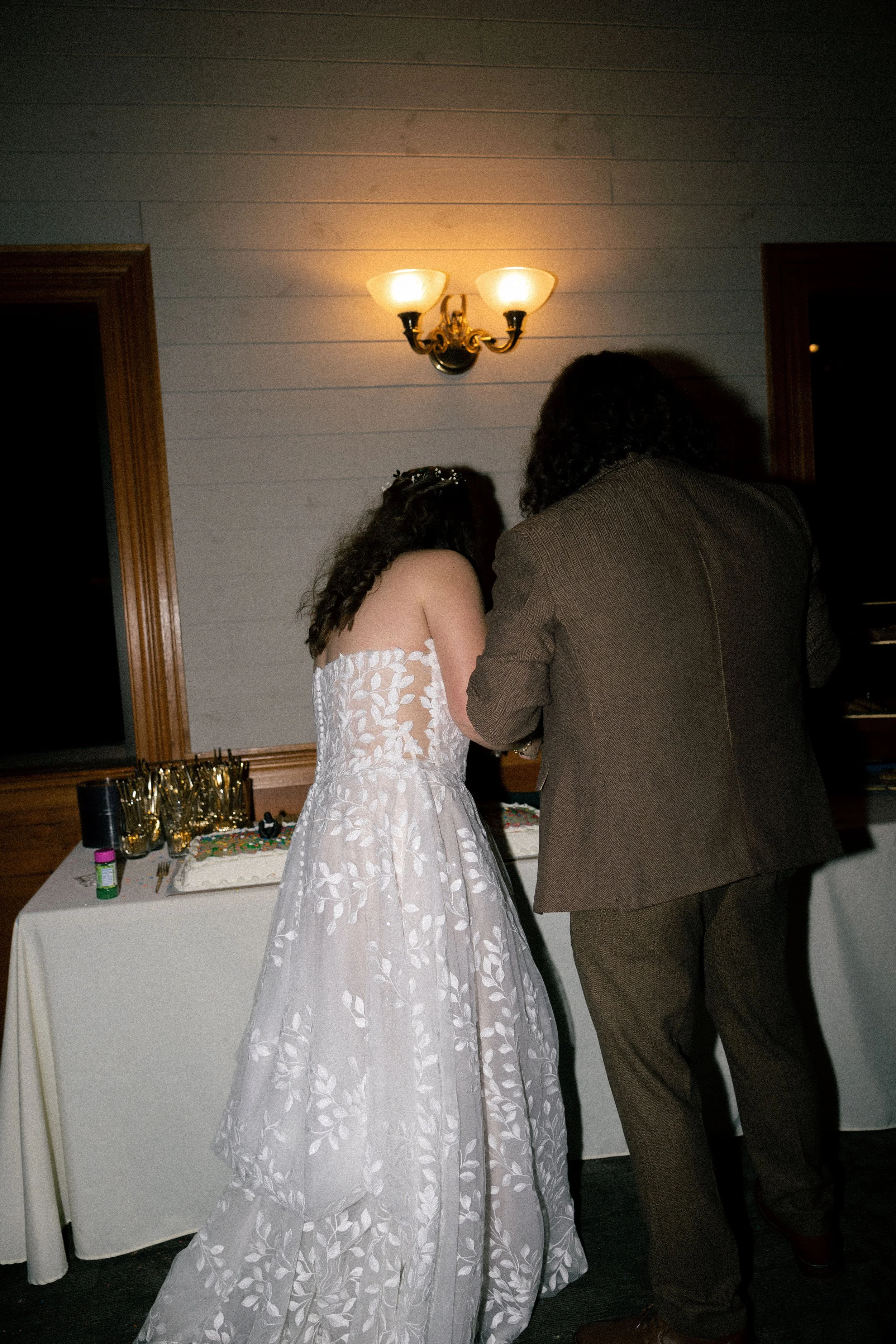 A bride and groom at a wedding reception cutting a cake together, with glasses and decorations on the table nearby, and a wall sconce lighting above them.