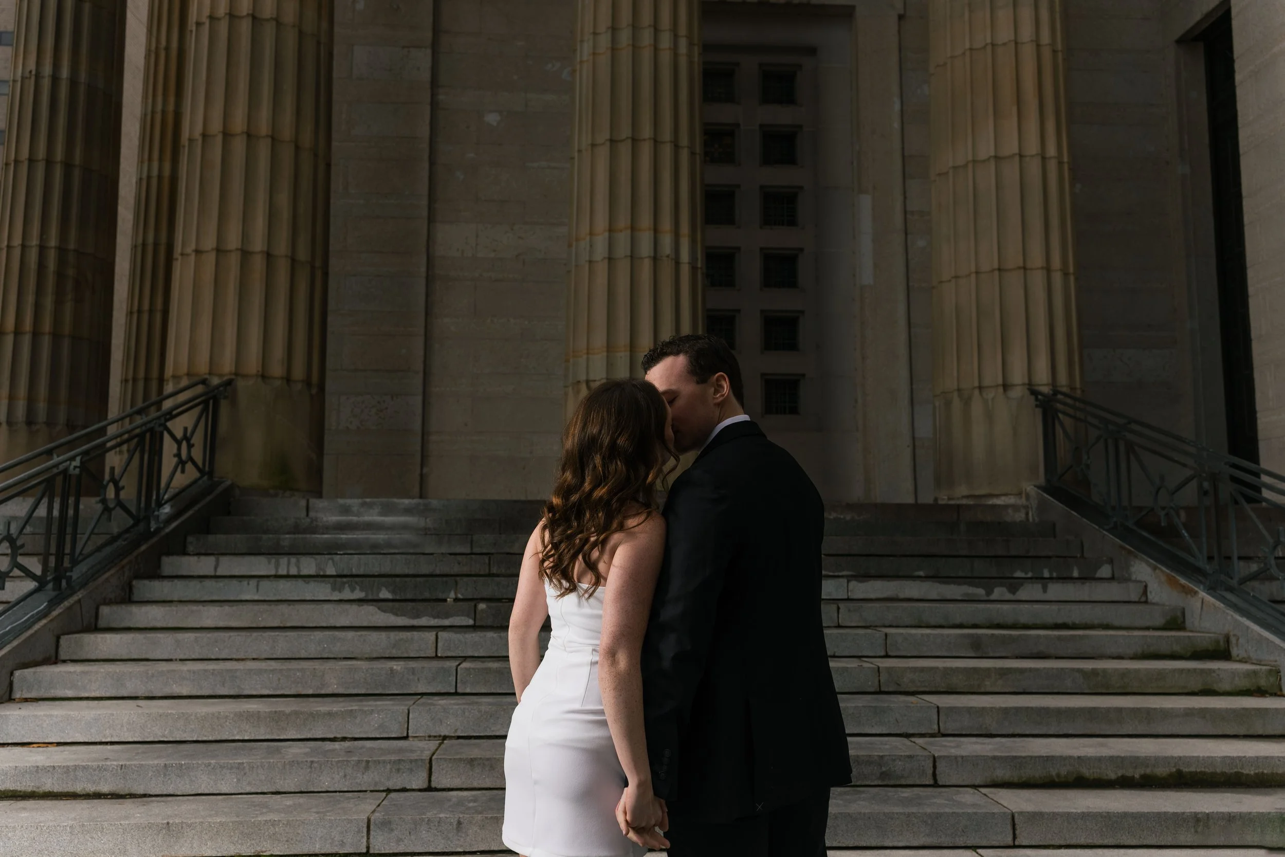 A couple in wedding attire, holding hands and kissing in front of a grand, classical building with large columns and stone stairs.