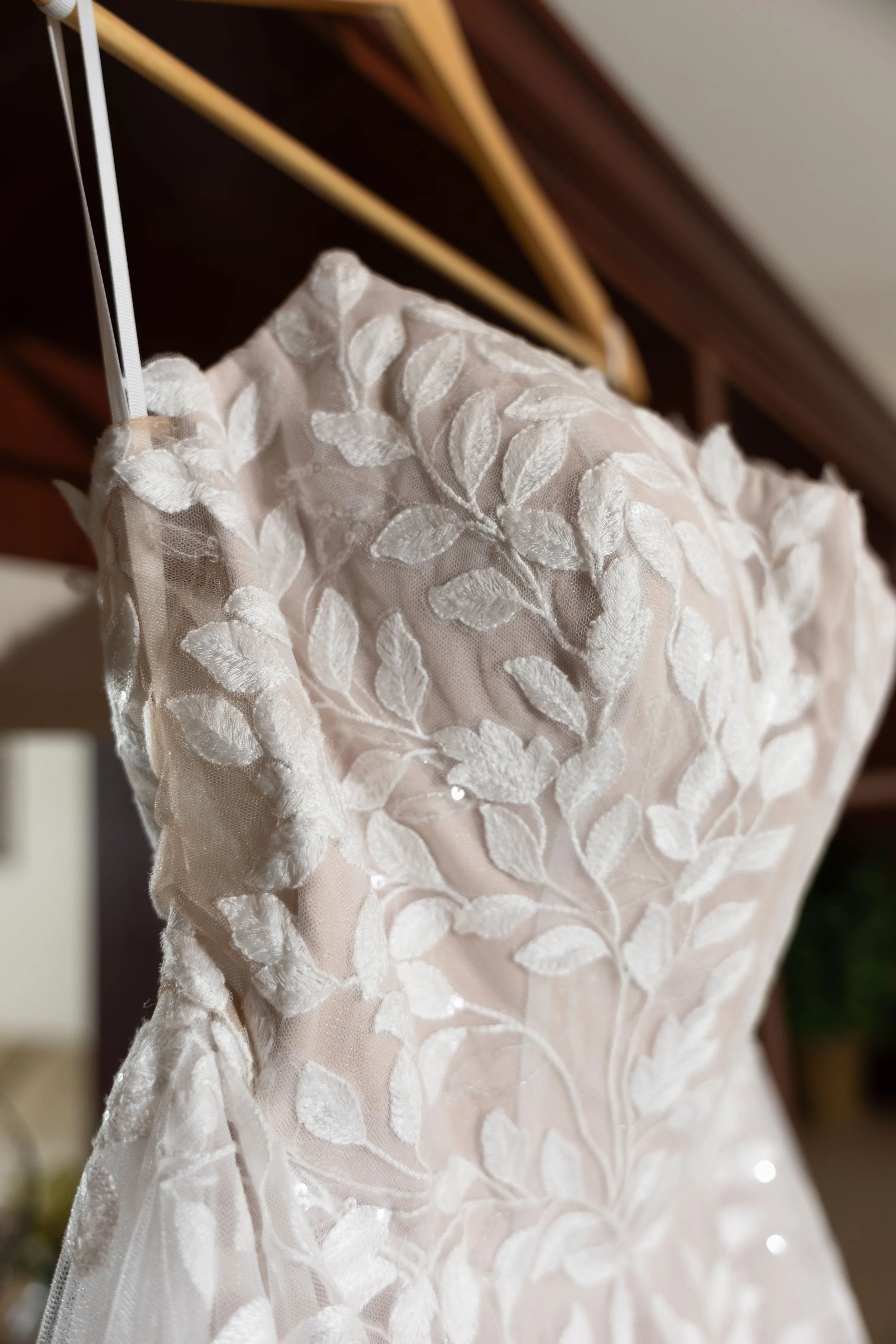 Close-up of a wedding dress with intricate white embroidery of leaves on sheer fabric, hanging on a wooden hanger.