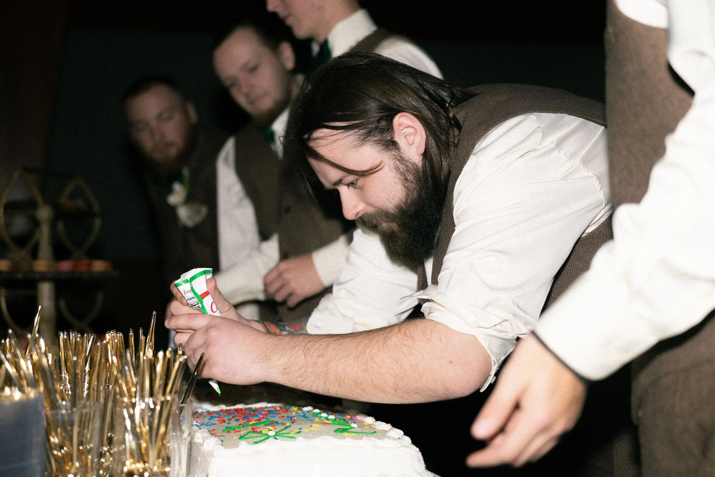A man with long dark hair and a beard decorating a white cake with colorful sprinkles at a gathering with other men in vests and dress shirts.