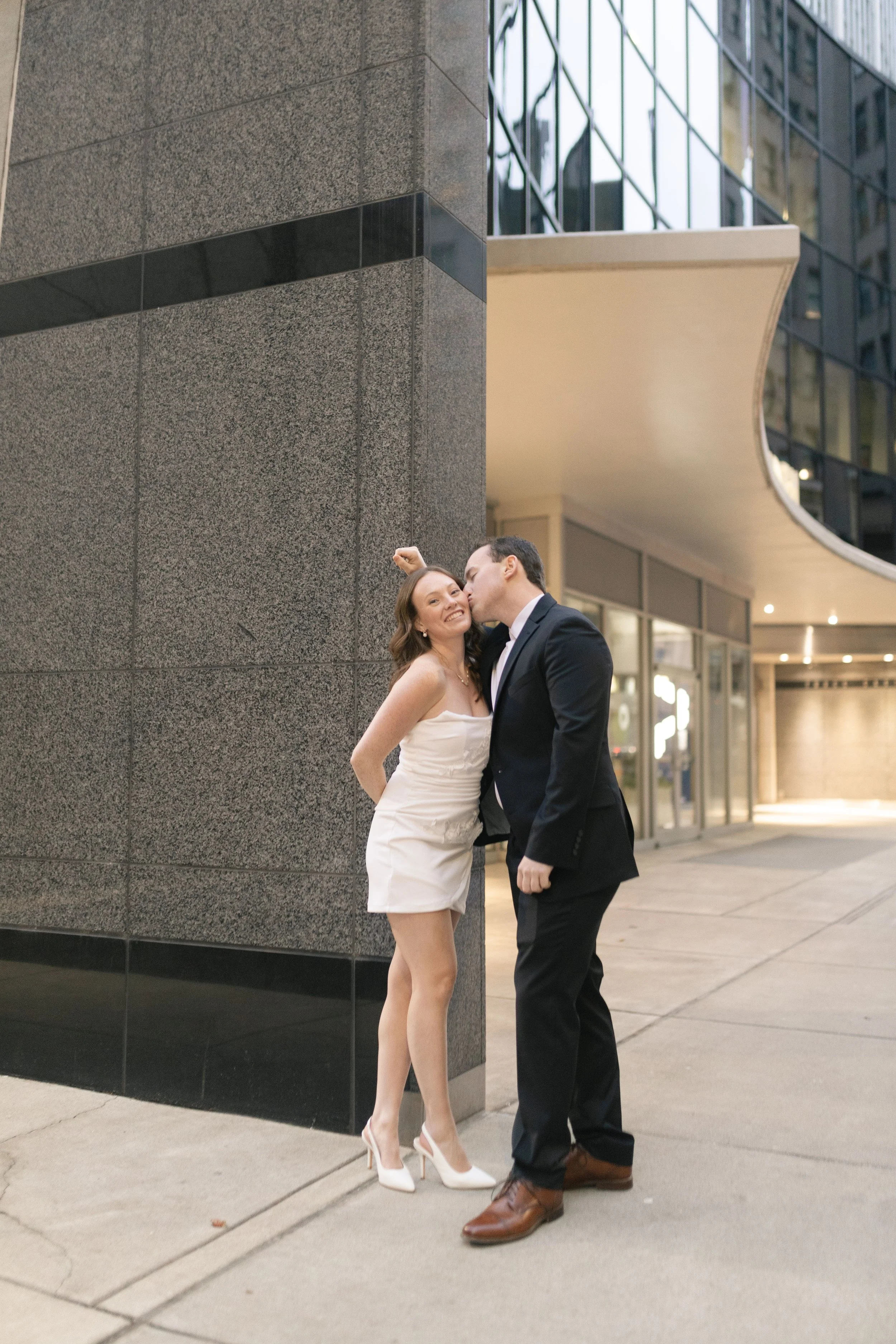 A young woman in a white dress and white heels standing on her toes while a man in a black suit and brown shoes kisses her cheek on an urban sidewalk at dusk.