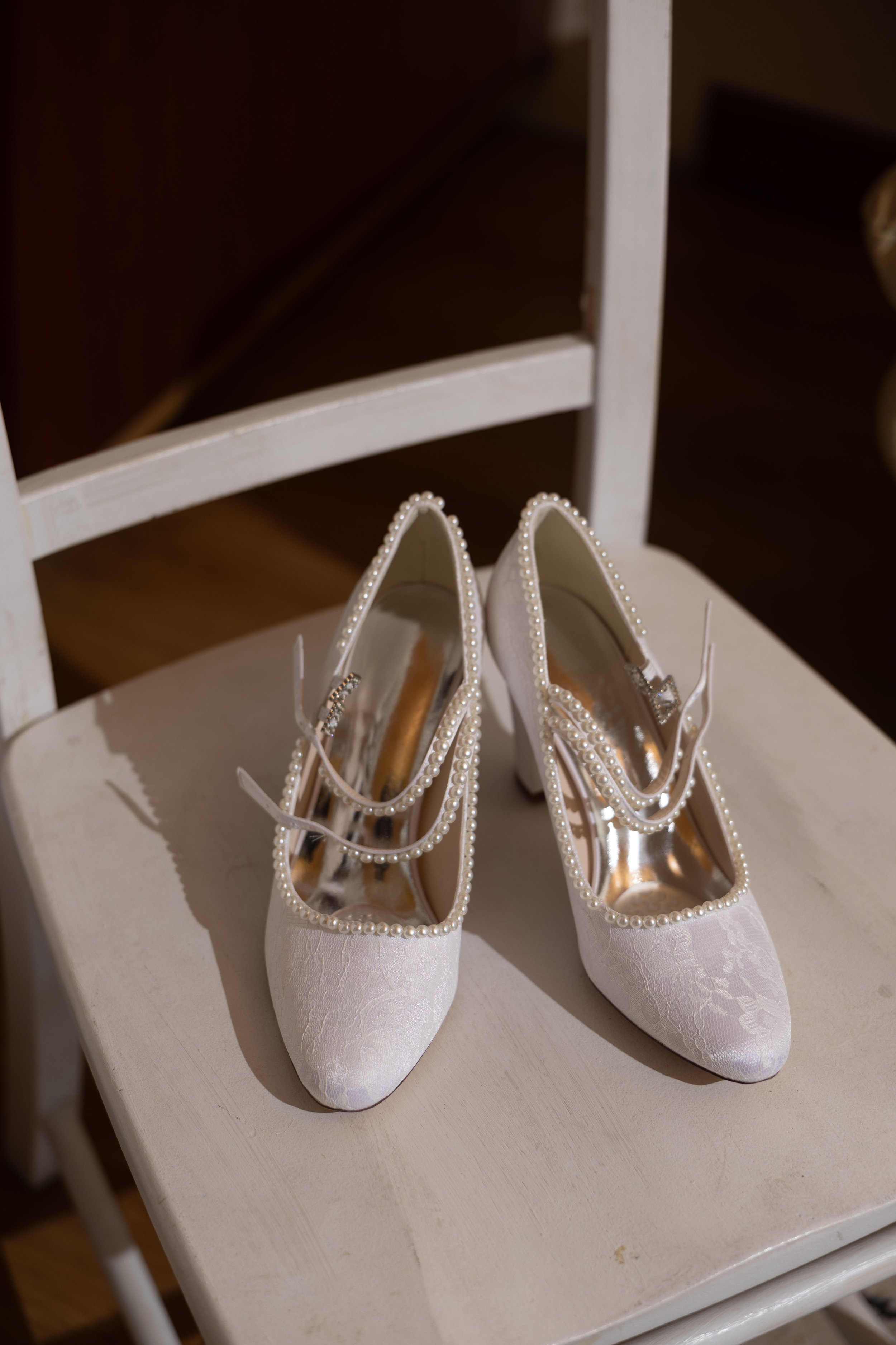 A pair of elegant white wedding shoes with pearl embellishments on a white chair.