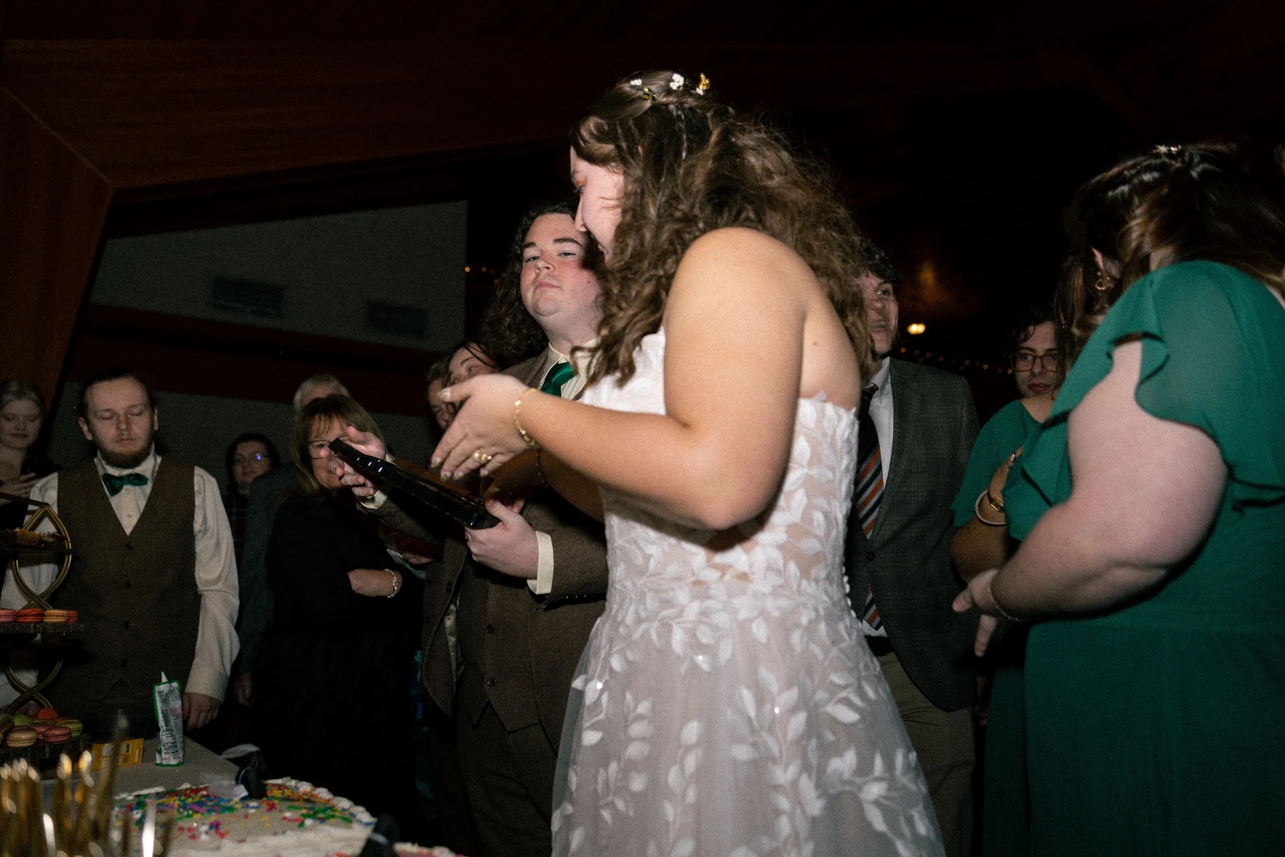 People gathered at a celebration with a woman in a white dress and a man holding a tray, near a cake with colorful sprinkles, in a dimly lit room.