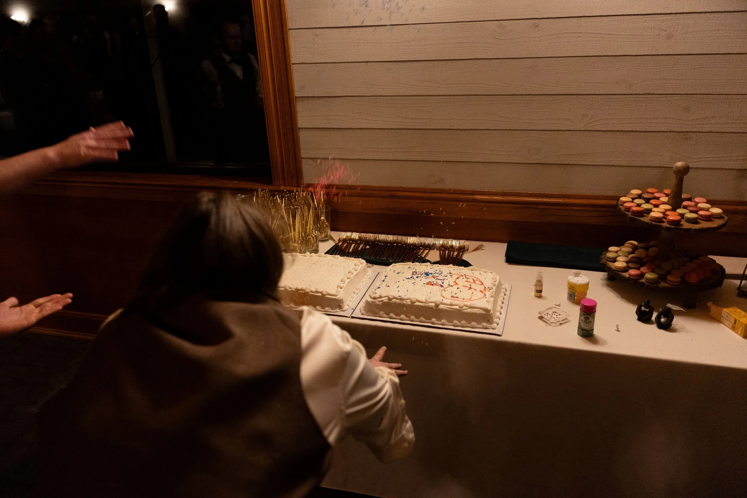 A person crouching in front of a table with two birthday cakes, one with a birthday message and another with colorful sprinkles, along with confetti and various party items.