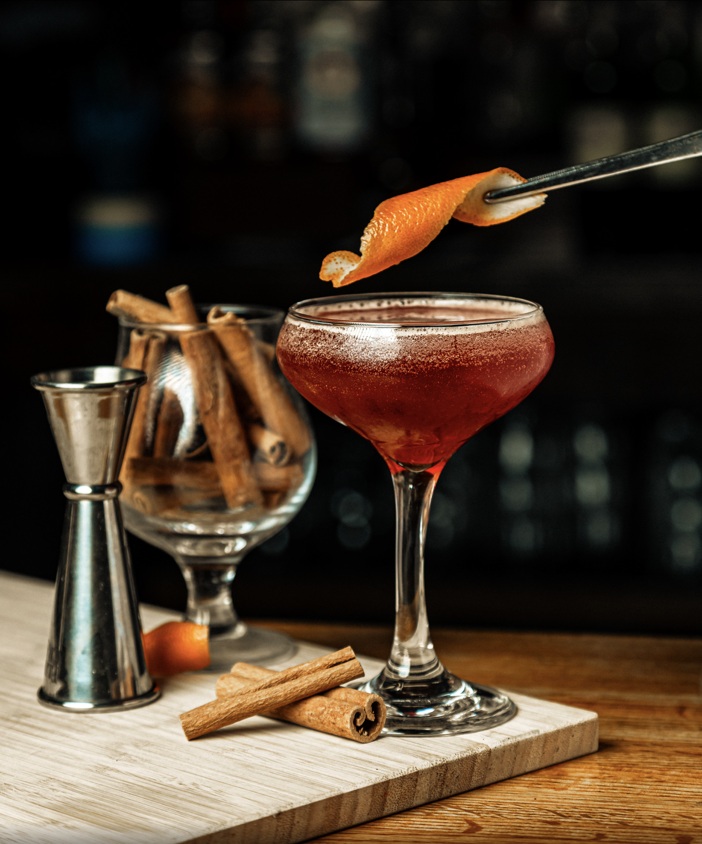 A red cocktail in a coupe glass with a slice of orange peel being added with tongs. There are cinnamon sticks, a lime wedge, and bartending tools on a wooden surface.