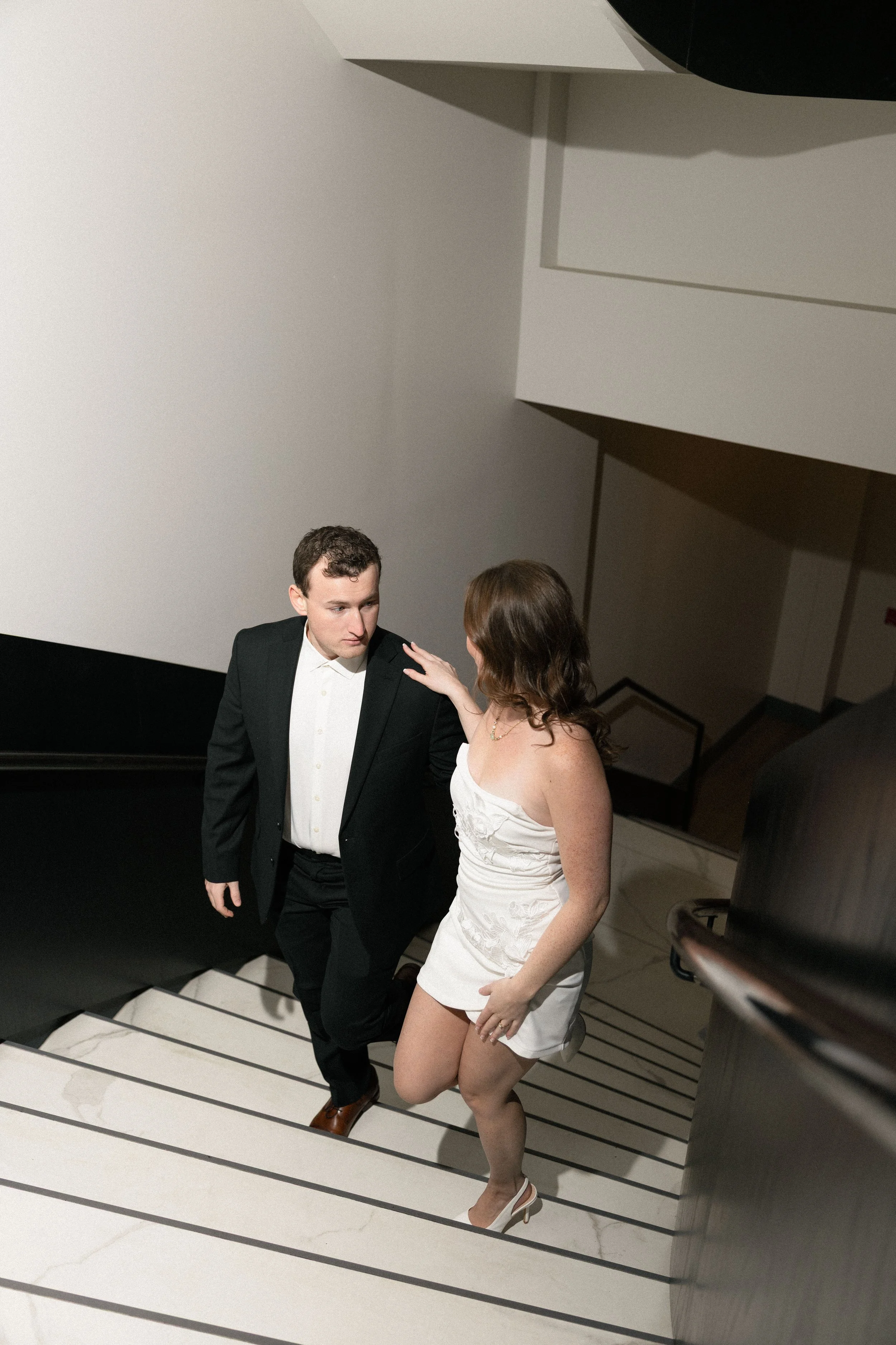 A man in a black suit and woman in a white dress walking up a staircase together, with the woman resting her hand on the man's shoulder.