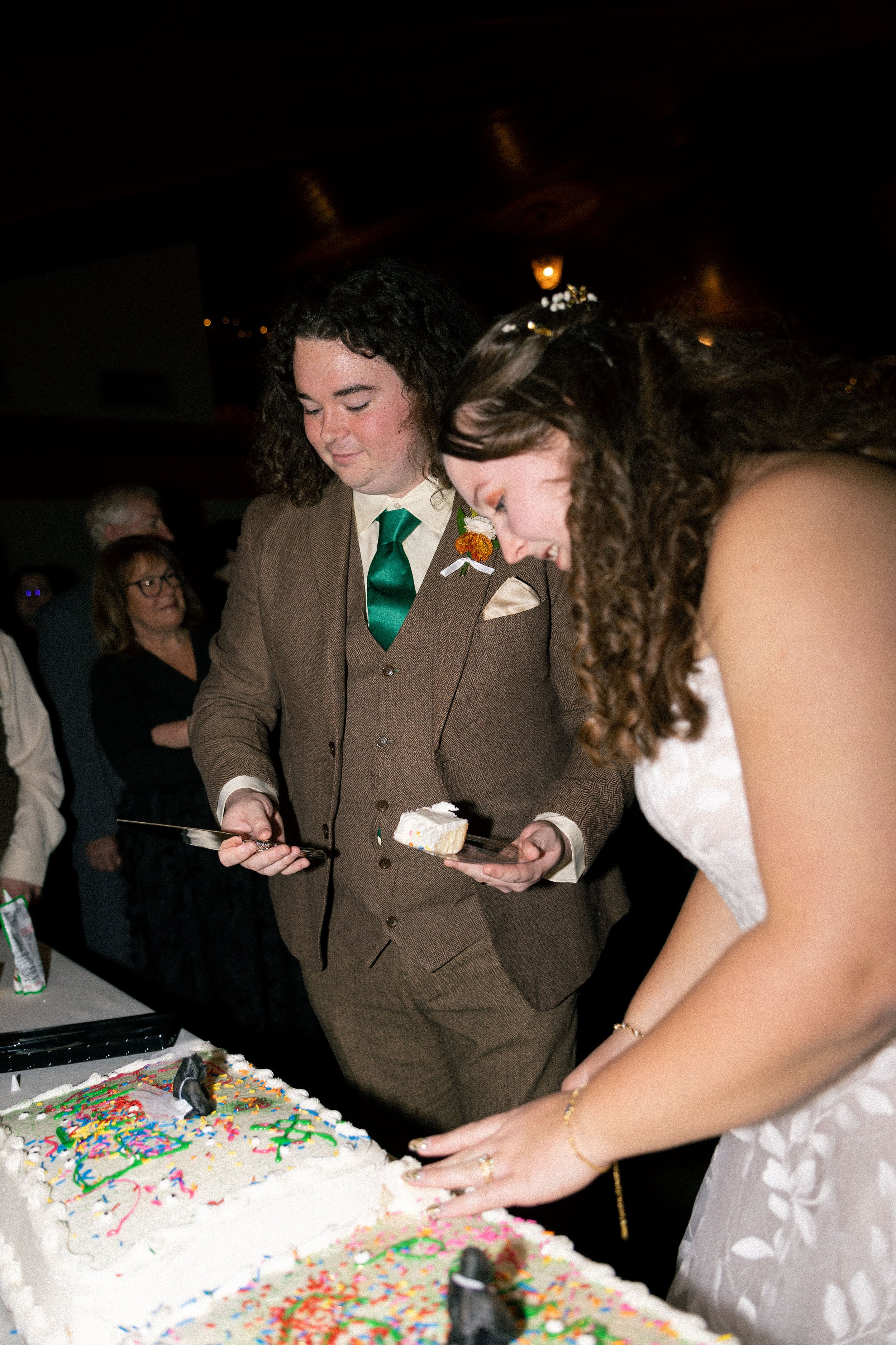 A couple at a wedding cake cutting ceremony. The groom is holding a knife and a plate with cake, while the bride is also cutting the cake. The cake is decorated with colorful sprinkles and topped with small decorations. Several guests are visible in 