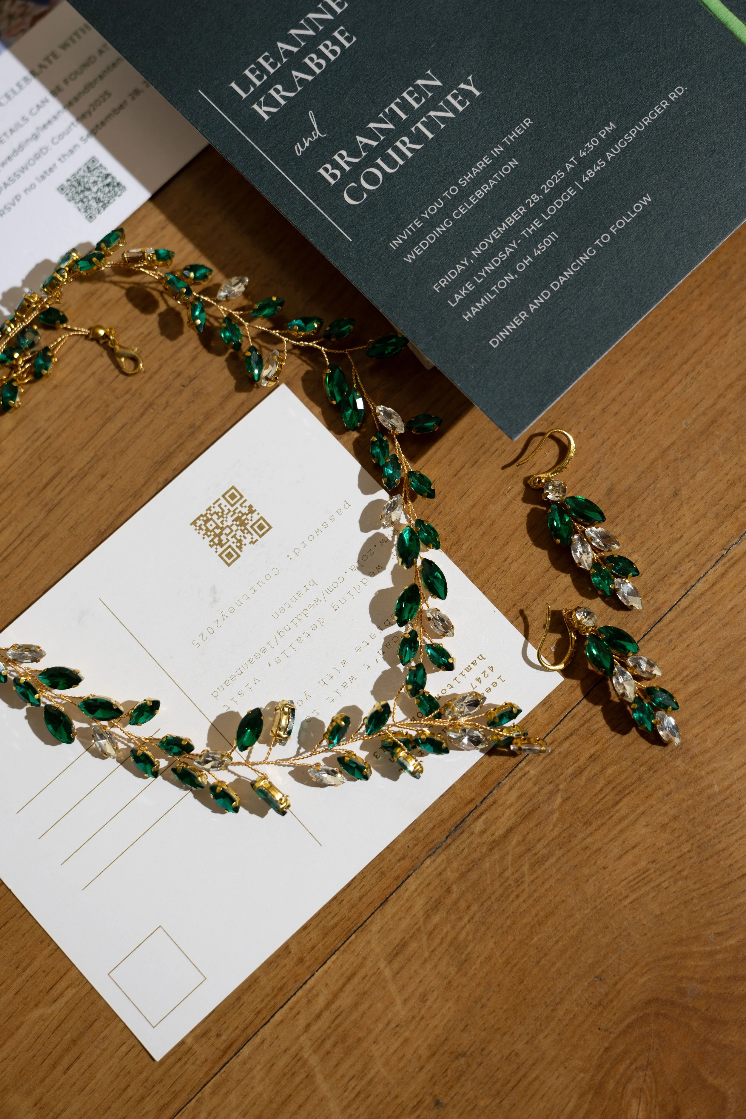 A wedding invitation, jewelry including a necklace and earrings with green and clear gemstones, and a white RSVP card with a QR code, all arranged on a wooden surface.