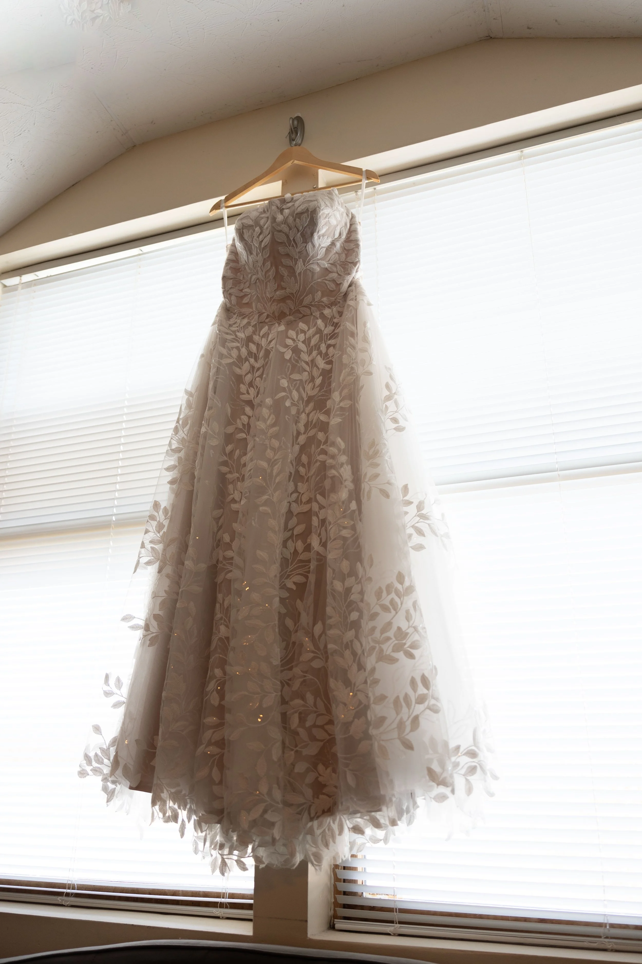A wedding dress hanging on a hanger in front of a window with blinds.