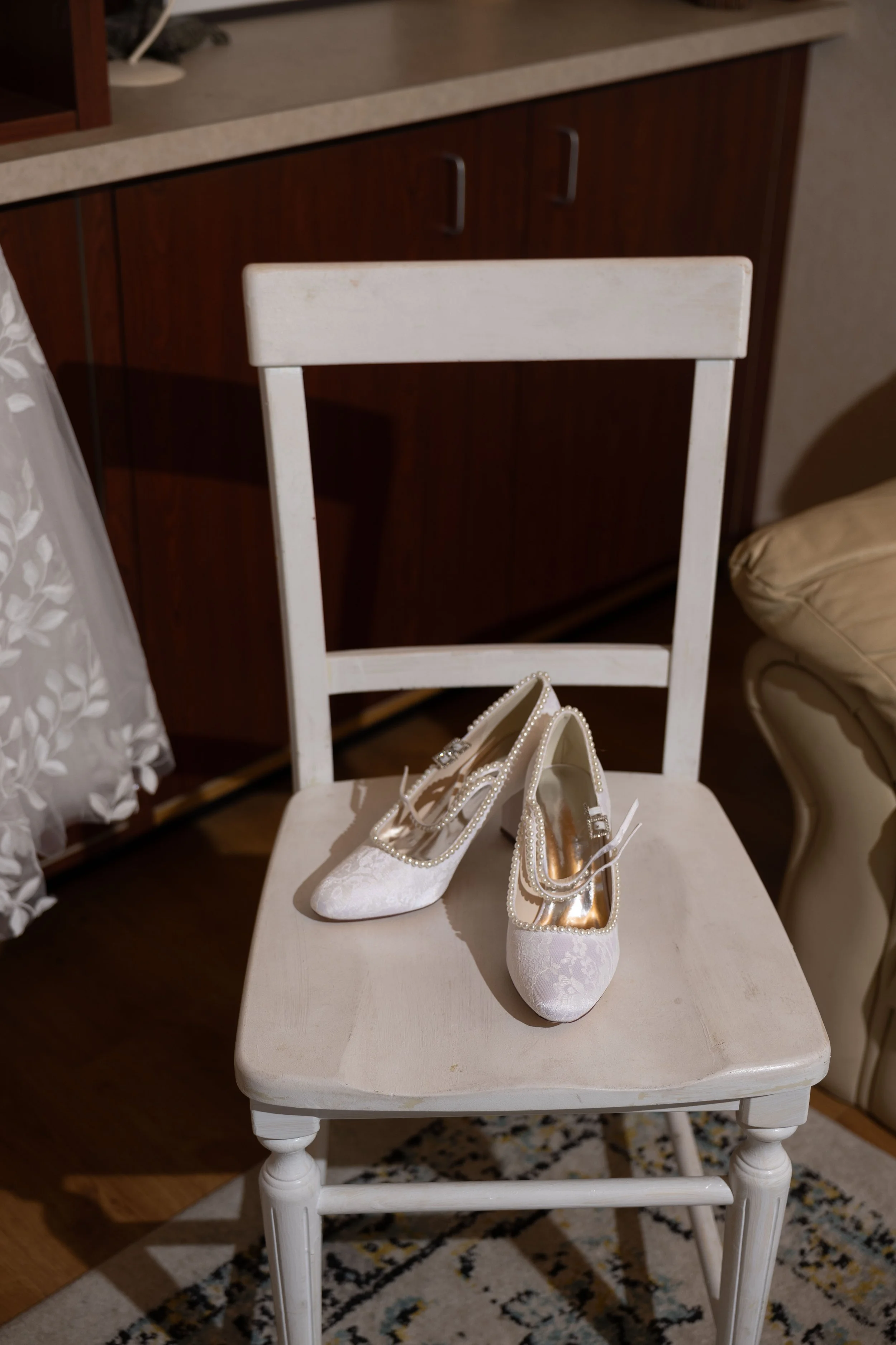 White bridal shoes with pearl decorations rest on a white wooden chair in a room with a patterned rug, a cream chair, and dark wooden cabinets.