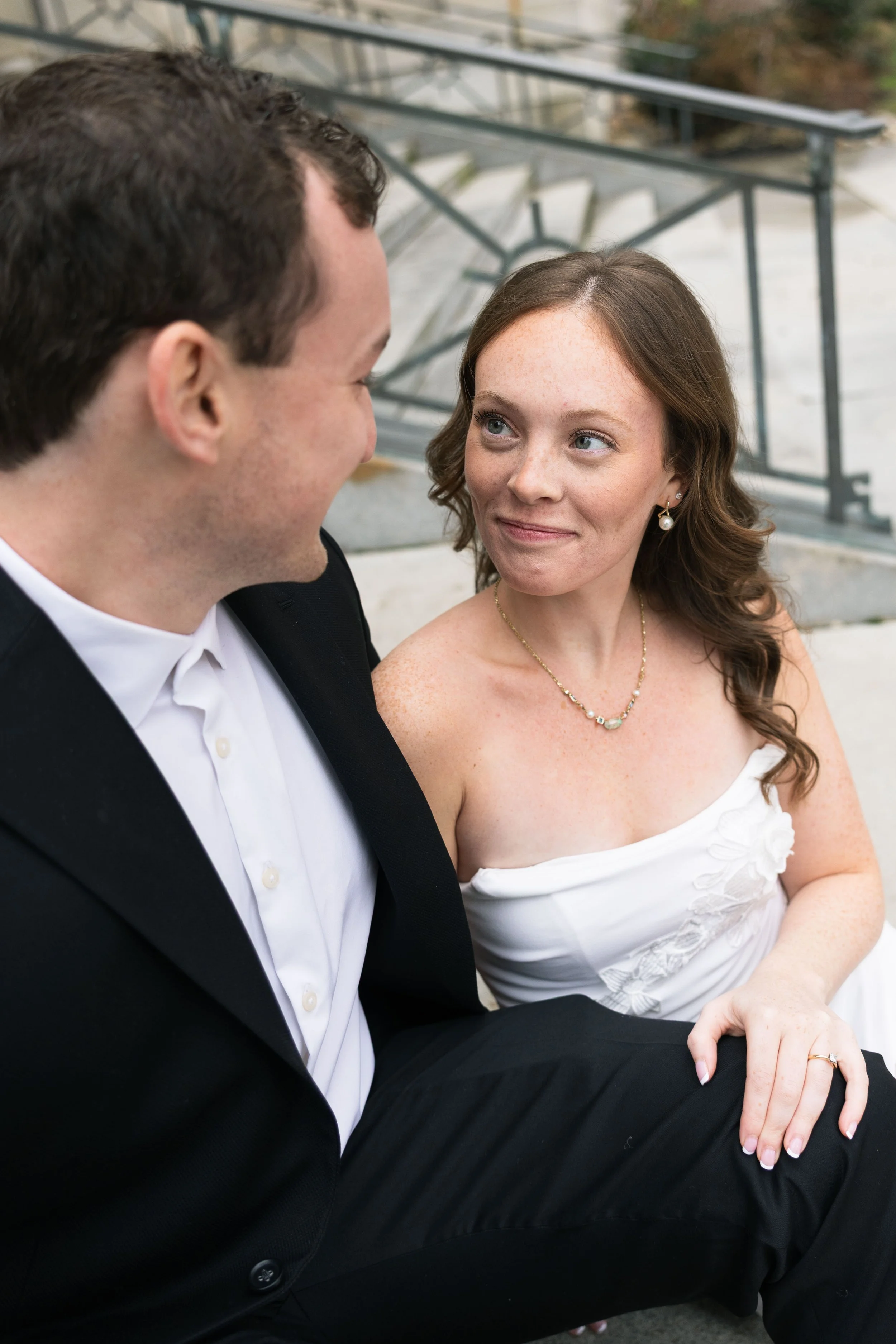 A bride and groom sitting on stairs outside, gazing into each other's eyes, smiling.