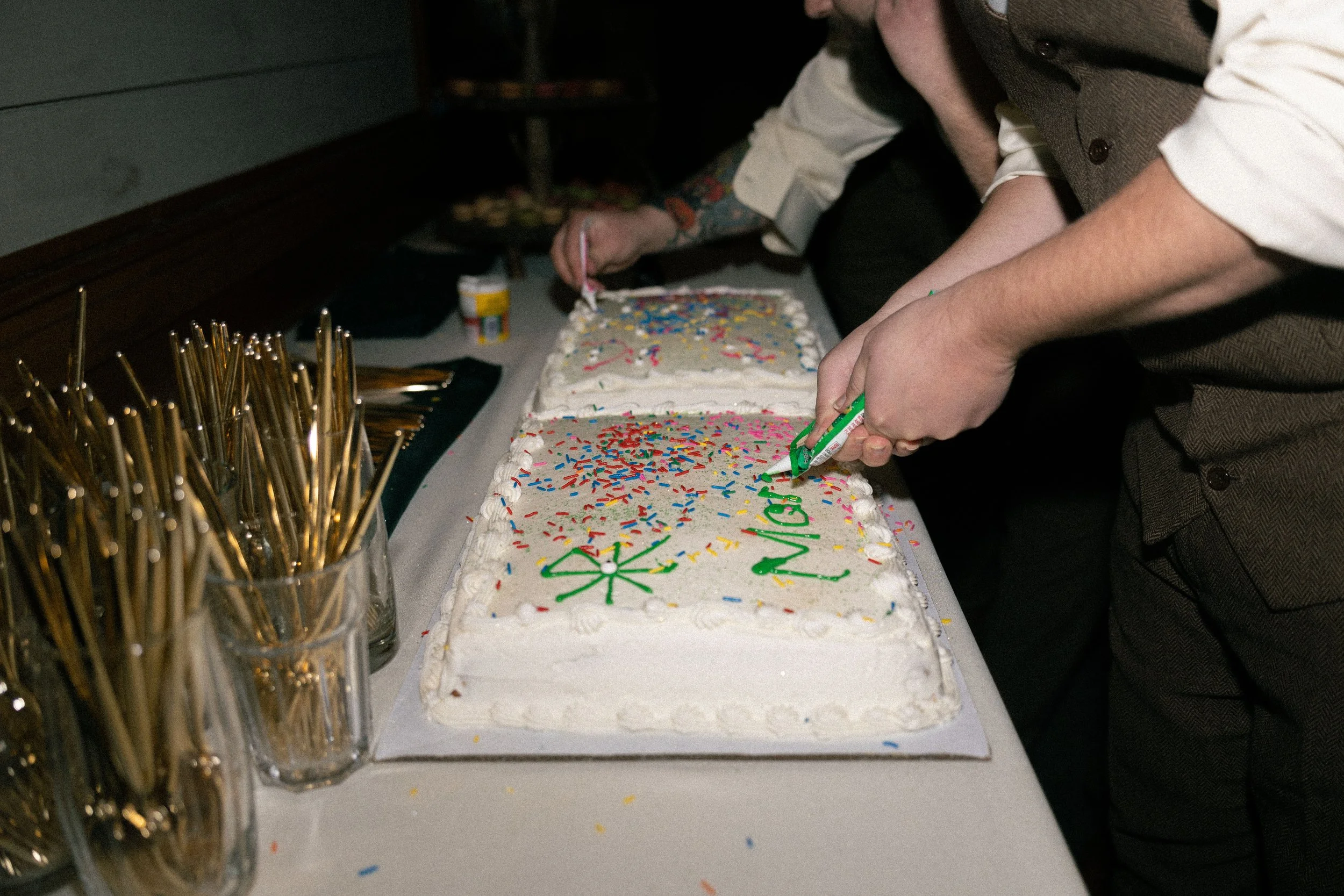 People gathered around a large rectangular birthday cake with white frosting, colorful sprinkles, and a message written in green and red icing. The cake is decorated with the words 'Happy Birthday' with candles and decorations.