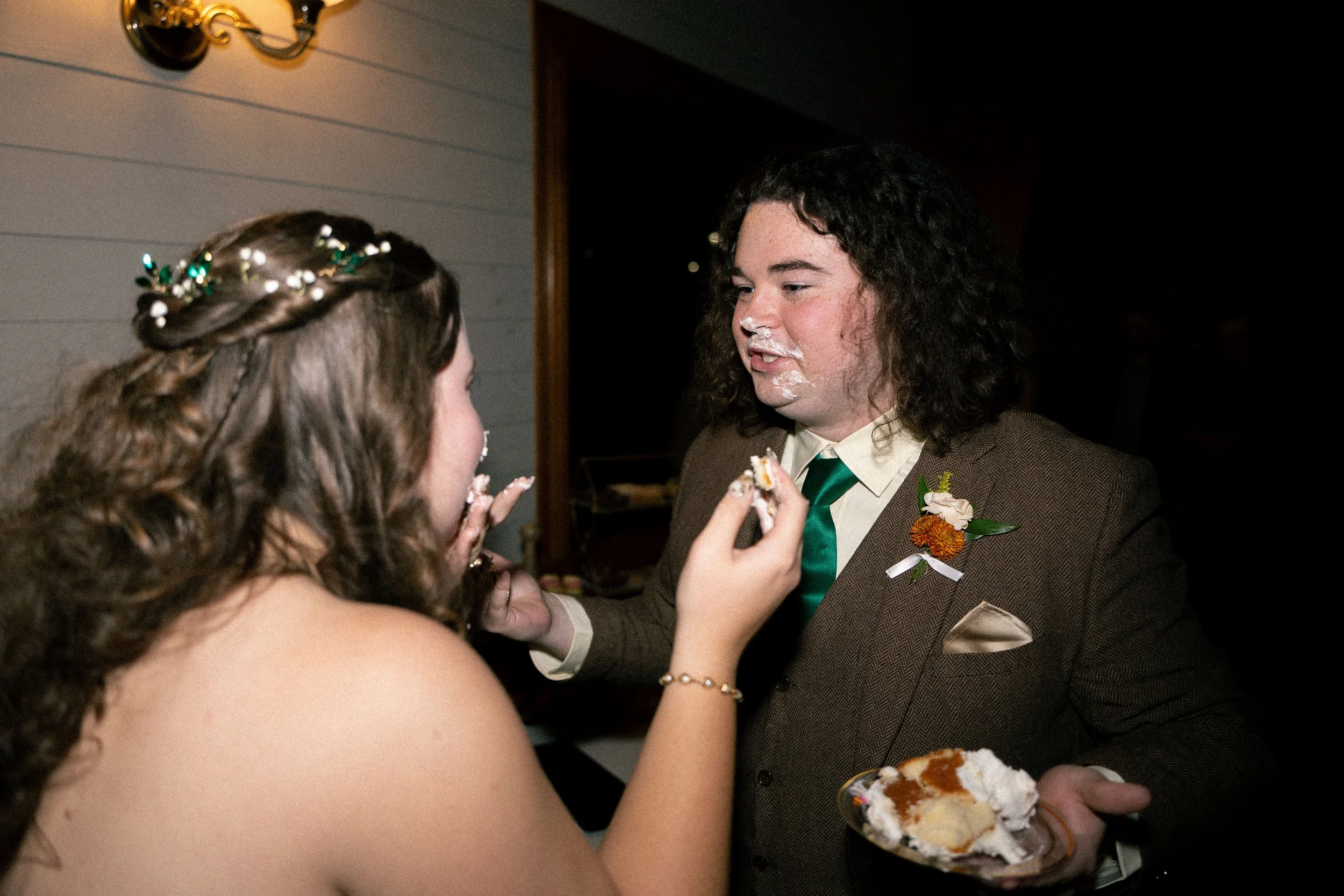 A couple, dressed in formal attire, are sharing cake at a wedding reception. The woman has long, curly hair with a decorative hairpiece, and the man has dark, curly hair and is wearing a suit with a boutonniere. The woman is feeding cake to the man, 