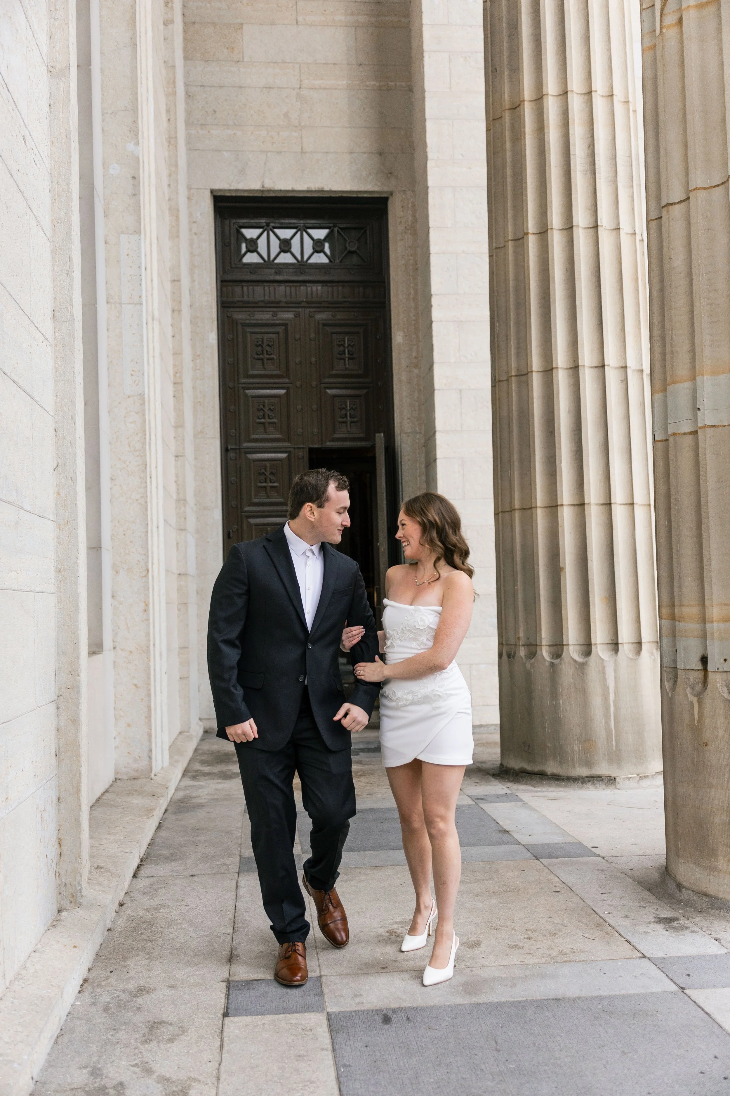 A couple walking arm in arm outside a building with large columns, smiling at each other.
