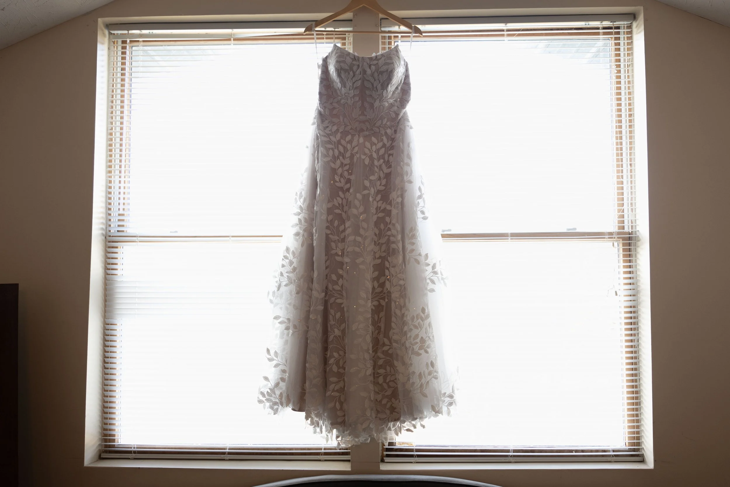White wedding dress hanging in front of a window with blinds, backlit sunlight.