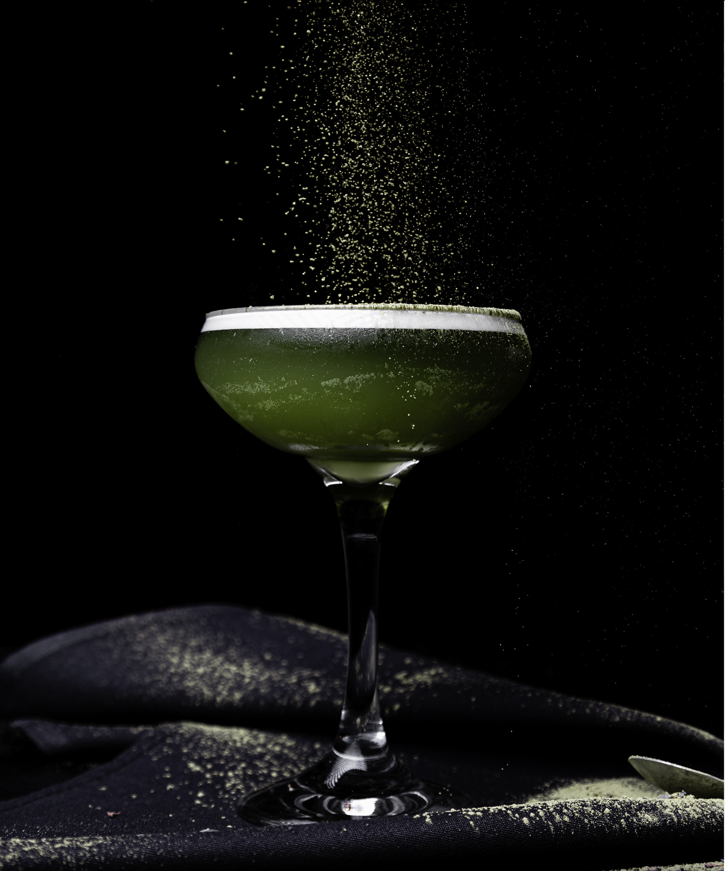 A cocktail glass filled with a green drink, topped with a white foam, with yellow powder sprinkled from above against a black background.