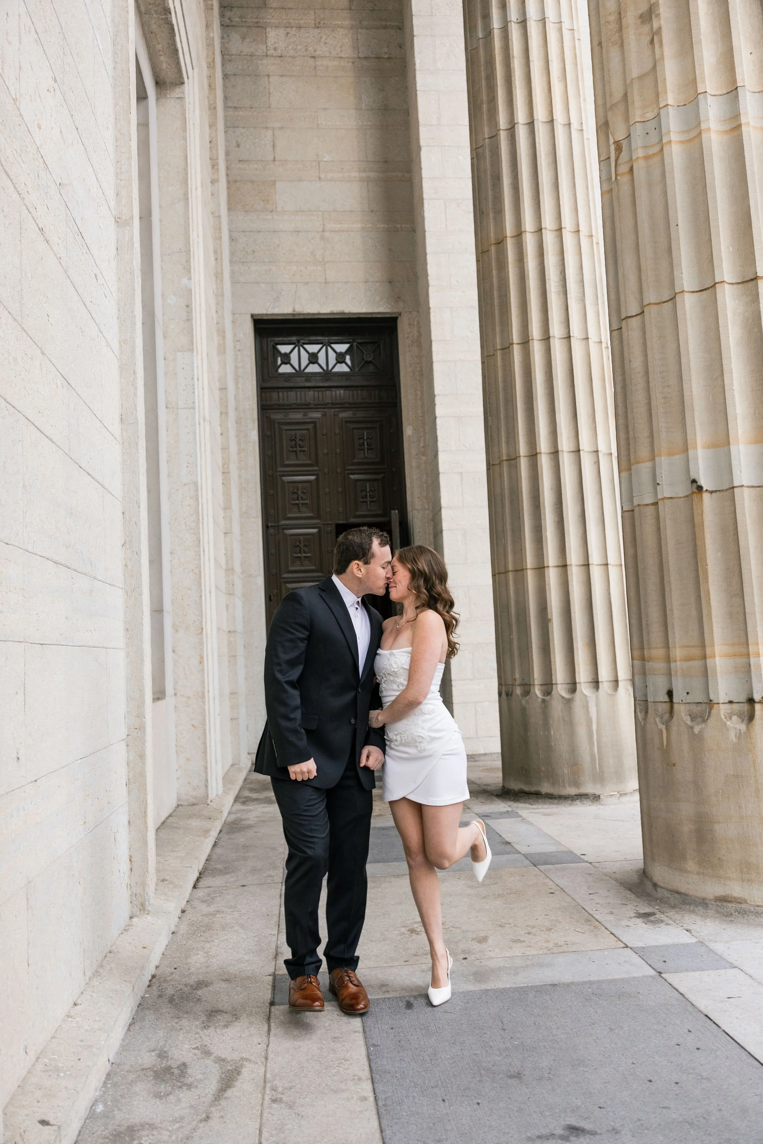 A couple dressed in wedding attire sharing a kiss in front of large stone columns and a wooden door, outdoors, during the day.