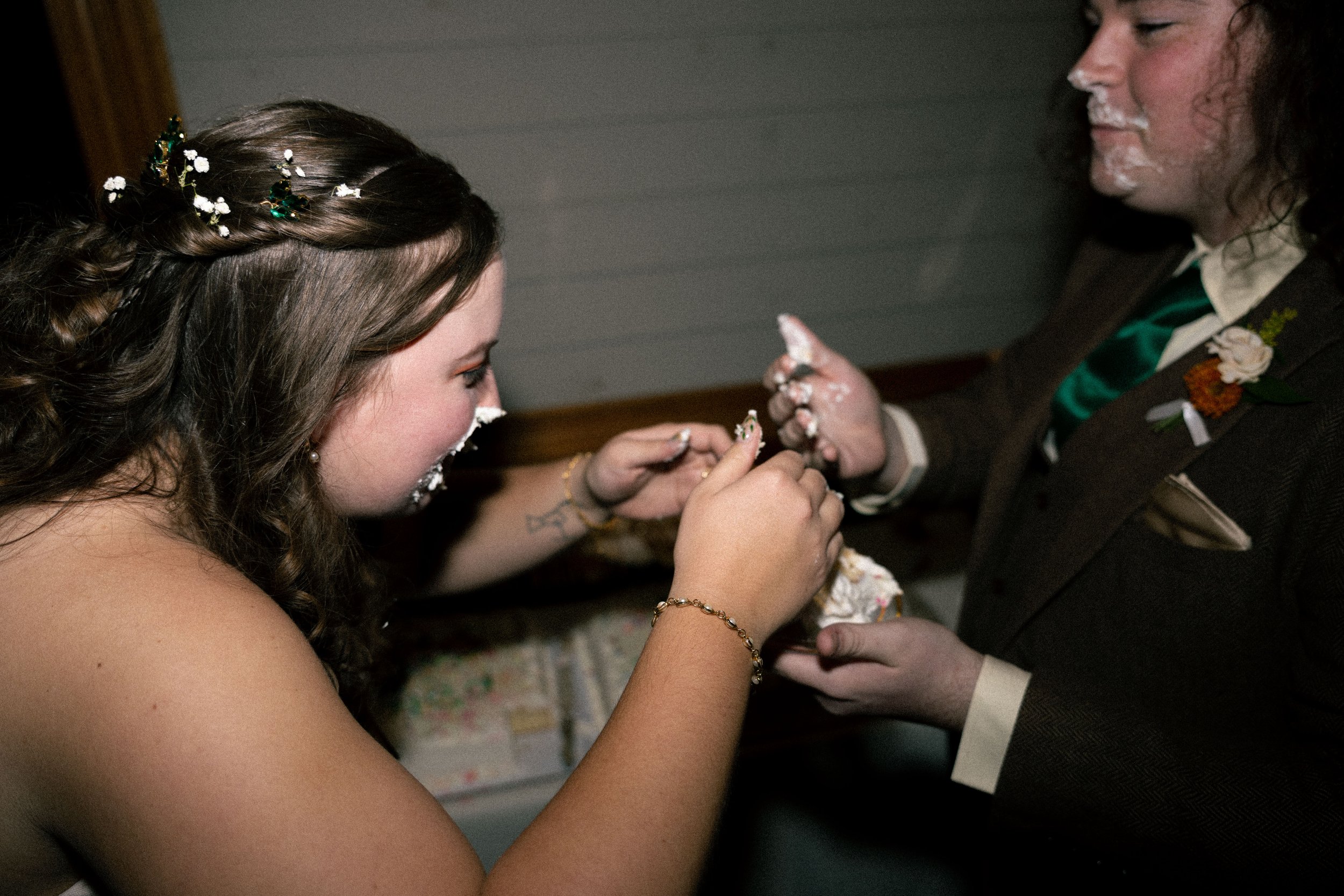 A bride with brown hair decorated with white flowers, smiling as she interacts with a groom during a wedding ceremony, with both having cake on their faces.