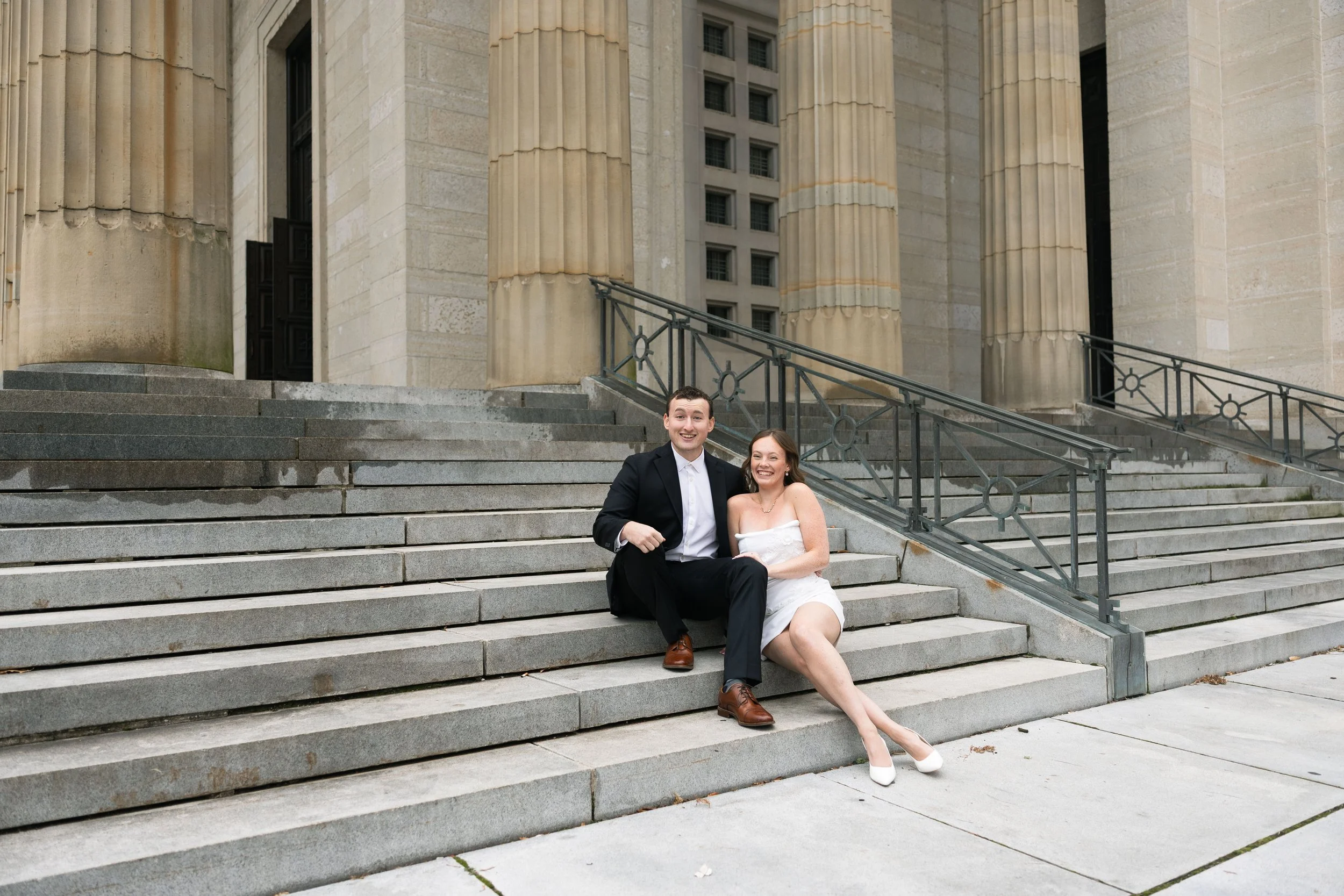 A man in a black tuxedo and a woman in a white dress sitting on the steps of a large, historic building with tall columns and a stone facade, smiling.