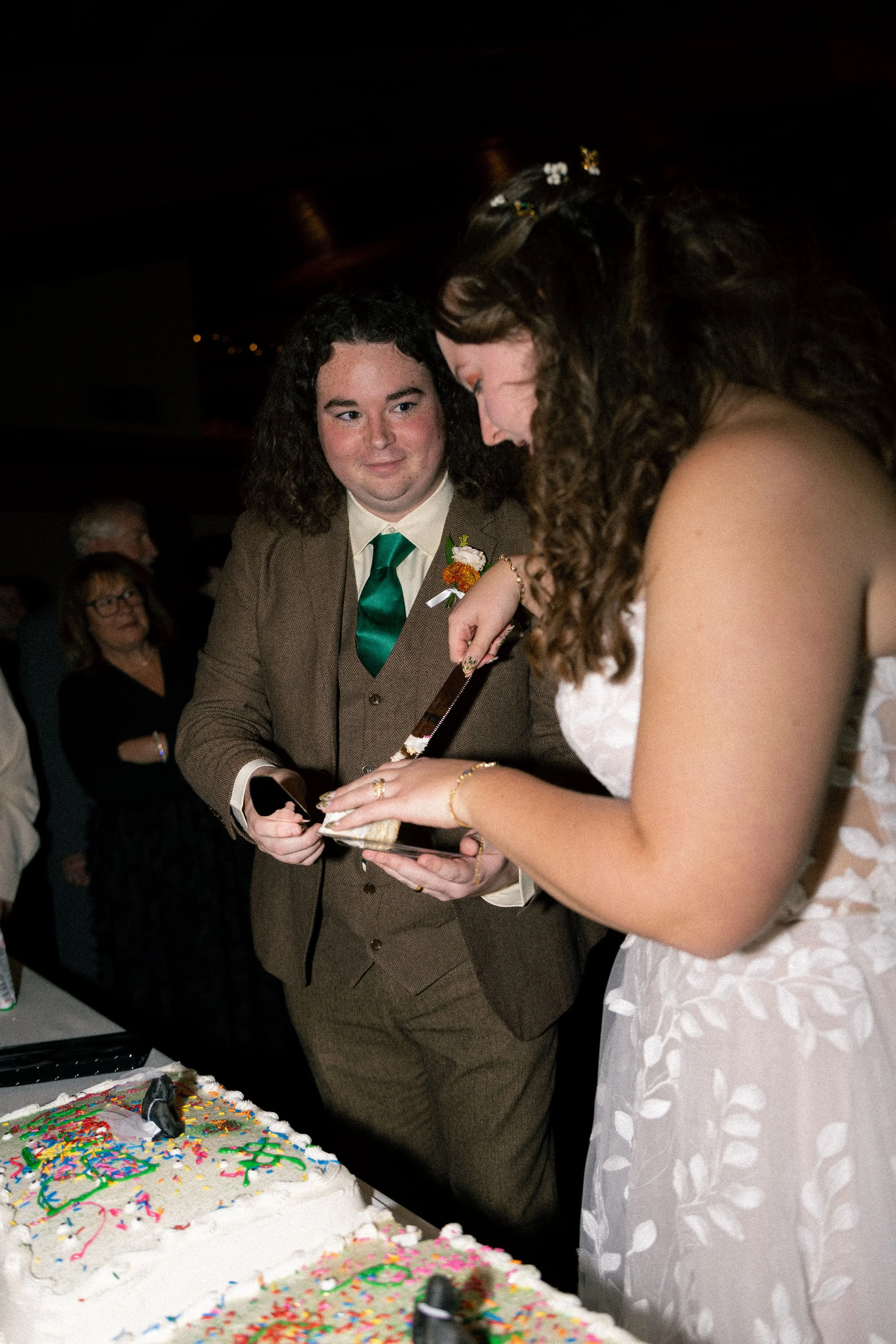 A couple dressed in wedding attire cutting a wedding cake together, surrounded by guests at a wedding reception.