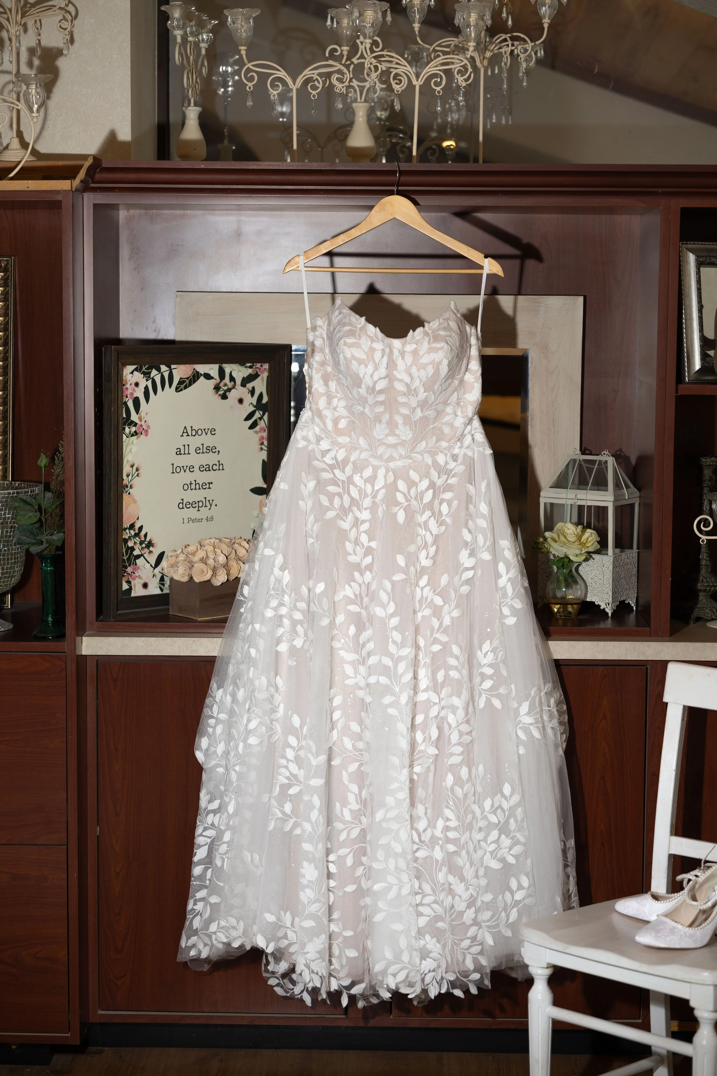 A white wedding dress with leaf embroidery hangs on a wooden hanger in a room with wooden furniture and decorative items, including a chandelier, framed quote, flowers, and a pair of white shoes on a chair.
