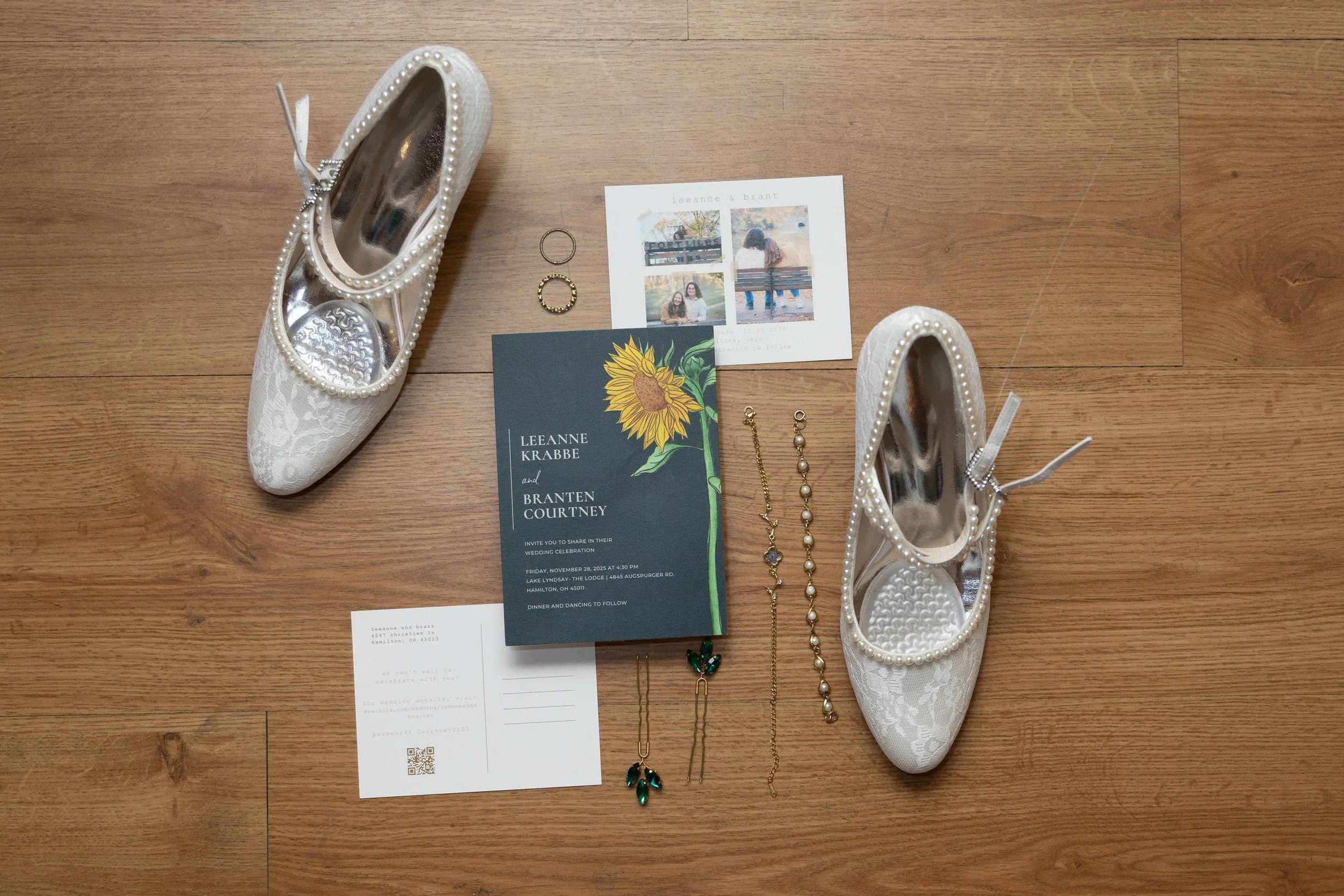 White lace wedding shoes with pearl embellishments, a wedding invitation, three rings, and assorted jewelry including earrings and necklaces, arranged on a wooden floor.