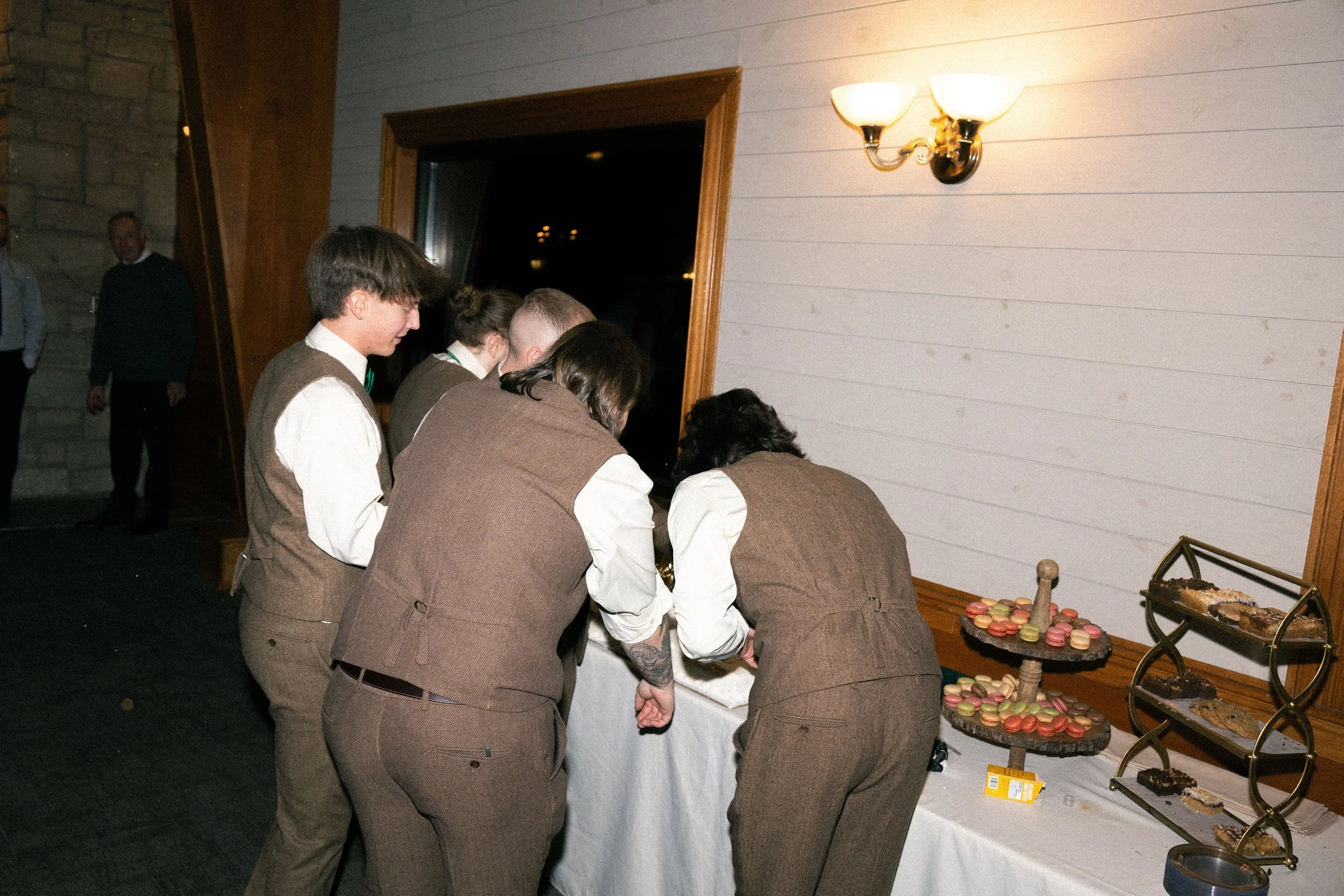 People in vintage-style clothing gathered around a table with desserts at a party or event.